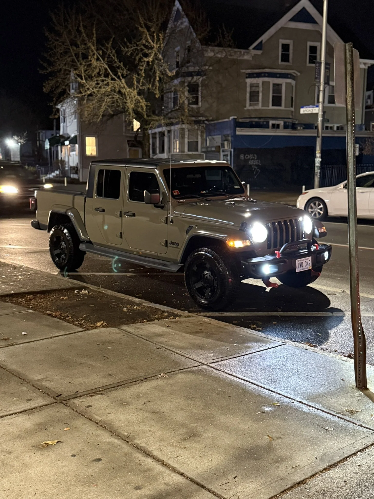 A beige Jeep Gladiator pickup truck parked on a city street at night, with its headlights on and a pole leaning to the right nearby.