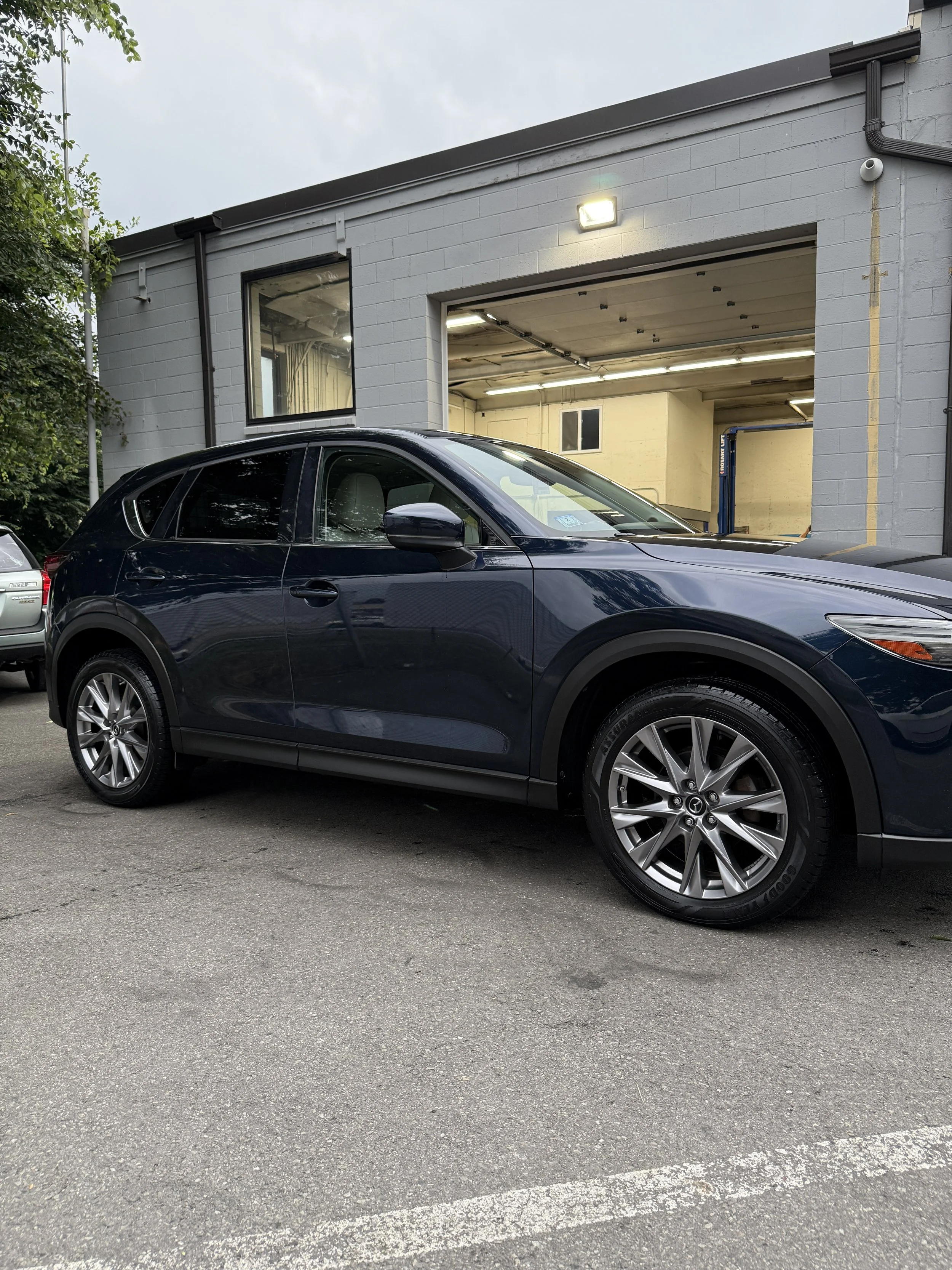 A blue Mazda SUV parked outside a gray building with a garage door open, revealing an interior with a lift and windows, in an overcast setting.