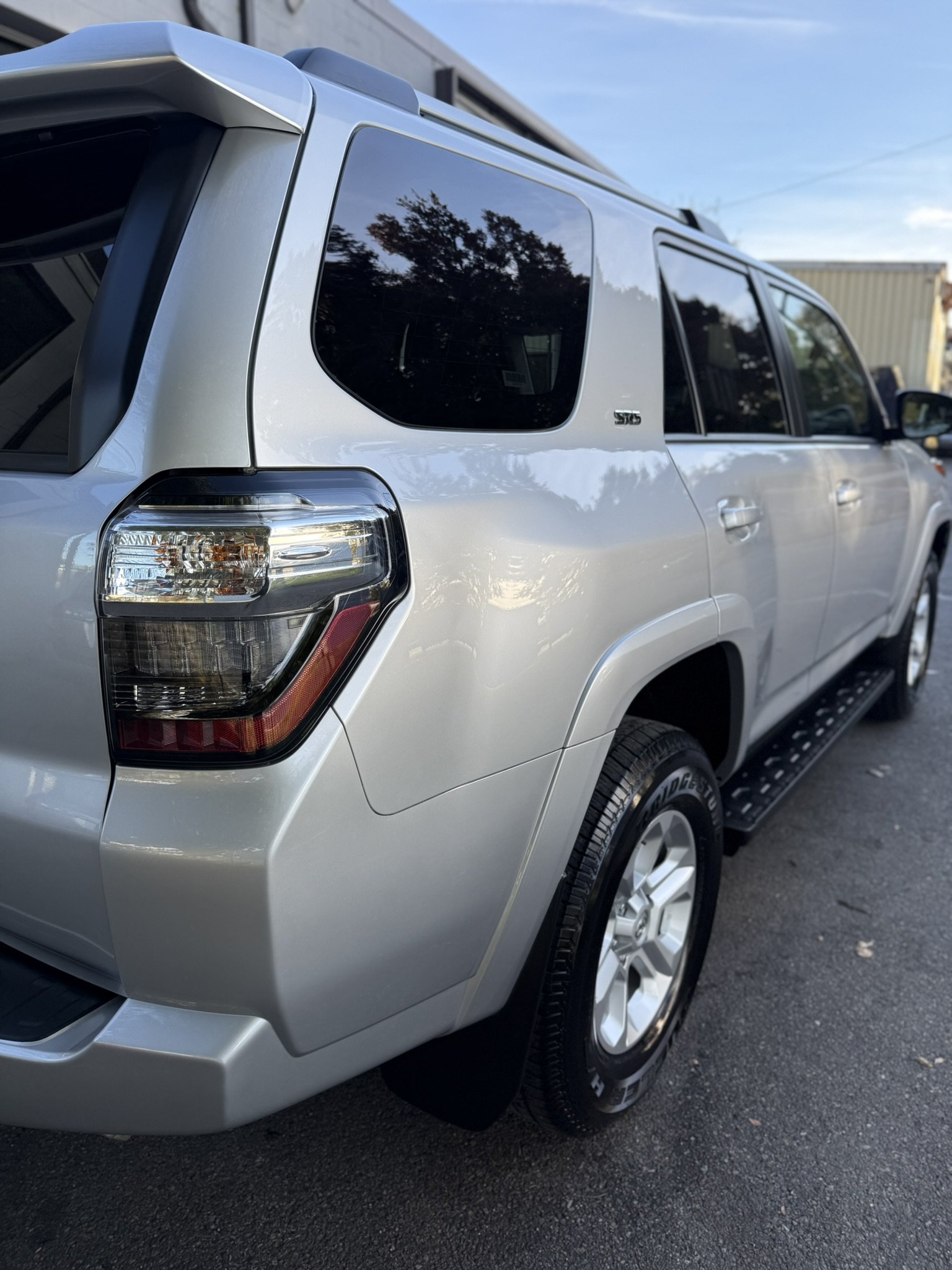 A silver Toyota 4Runner SUV parked outdoors on asphalt with a building and trees reflected on the windows, and a clear blue sky overhead.