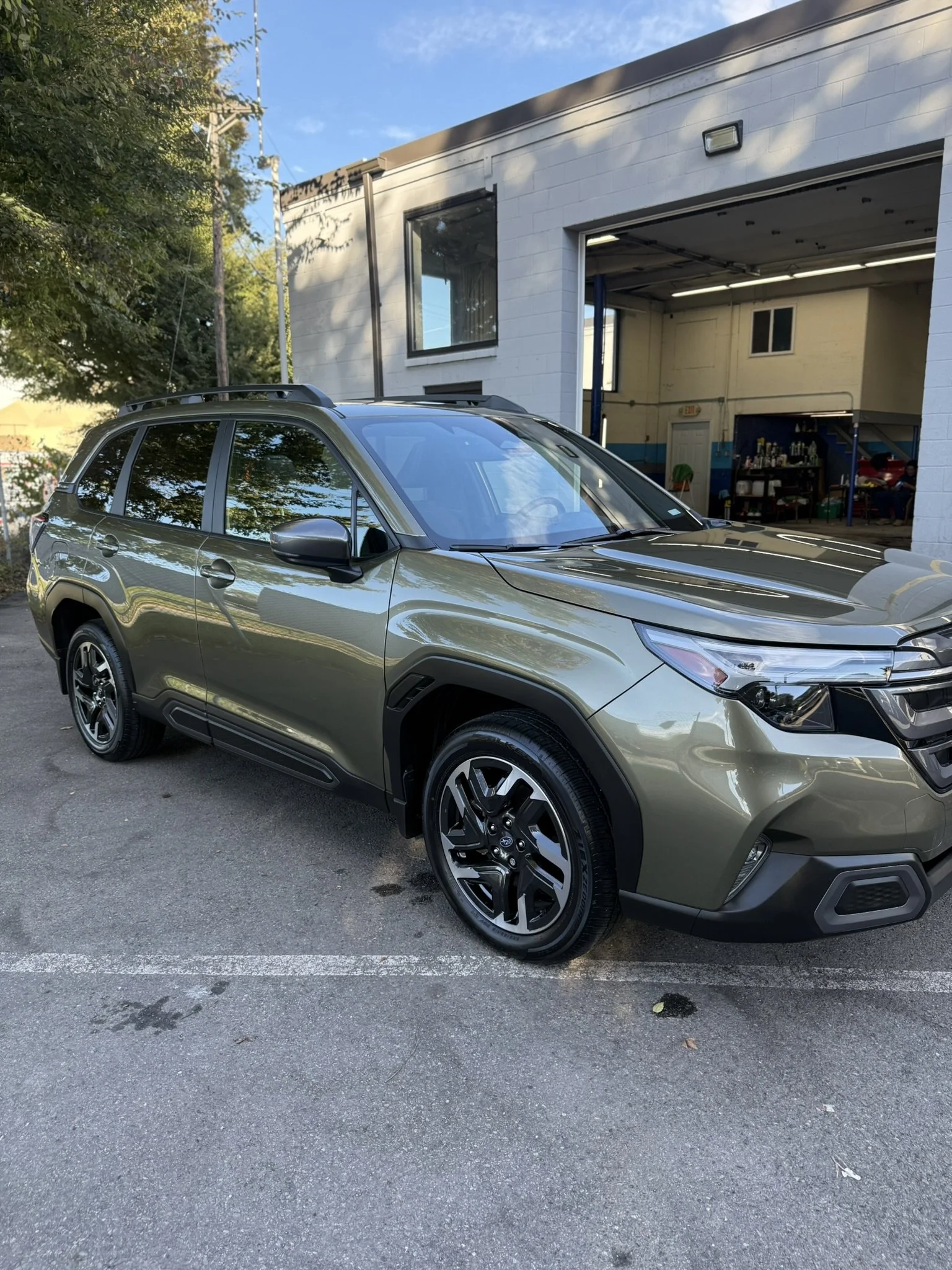 A green SUV parked in front of a building with an open garage door, reflecting trees on its surface, under a partly cloudy sky.