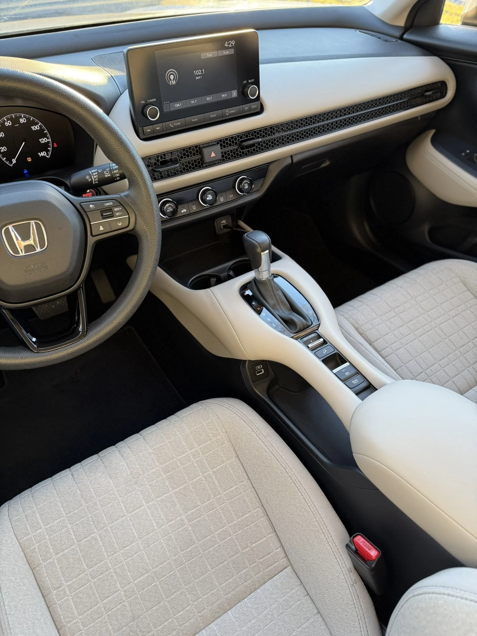 Interior of a Honda vehicle showing the steering wheel, dashboard with media screen, center console with gear shift, and beige cloth seats.