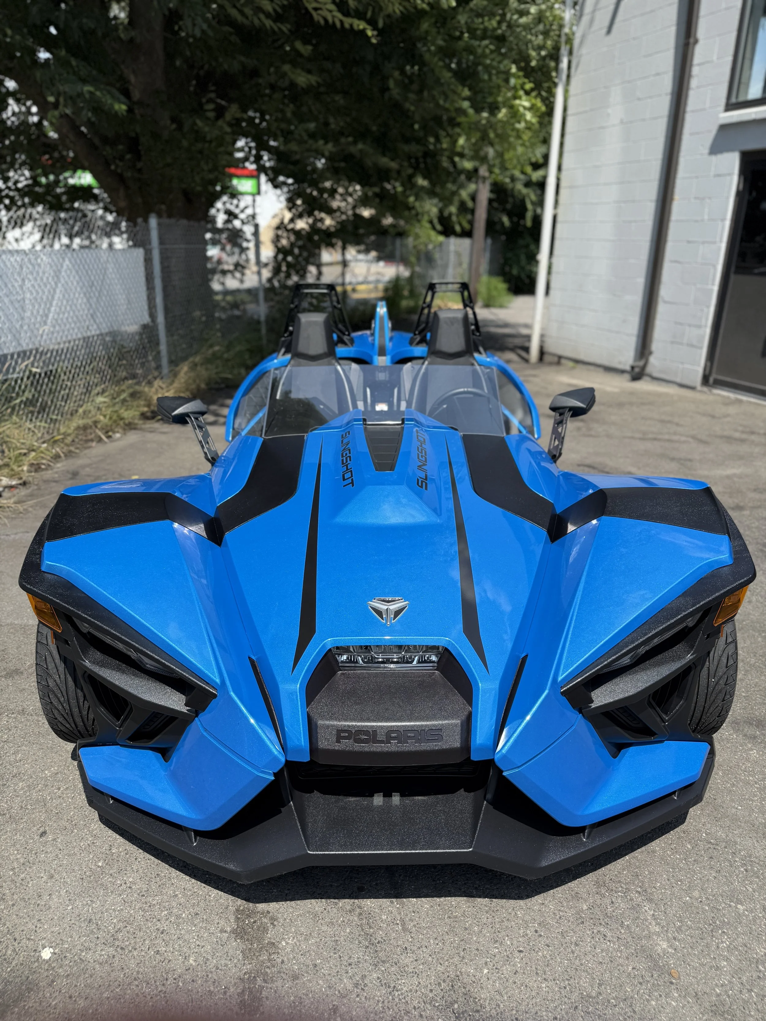 Blue Polaris Slingshot sports car parked on a concrete surface, with a white building on the right and trees in the background.