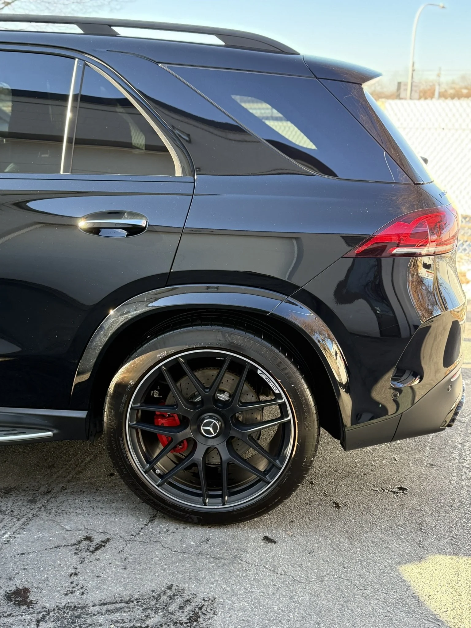 Close-up of the rear side of a black Mercedes-Benz SUV with AMG wheels and red brake calipers, parked on pavement near a fence and street lamp.
