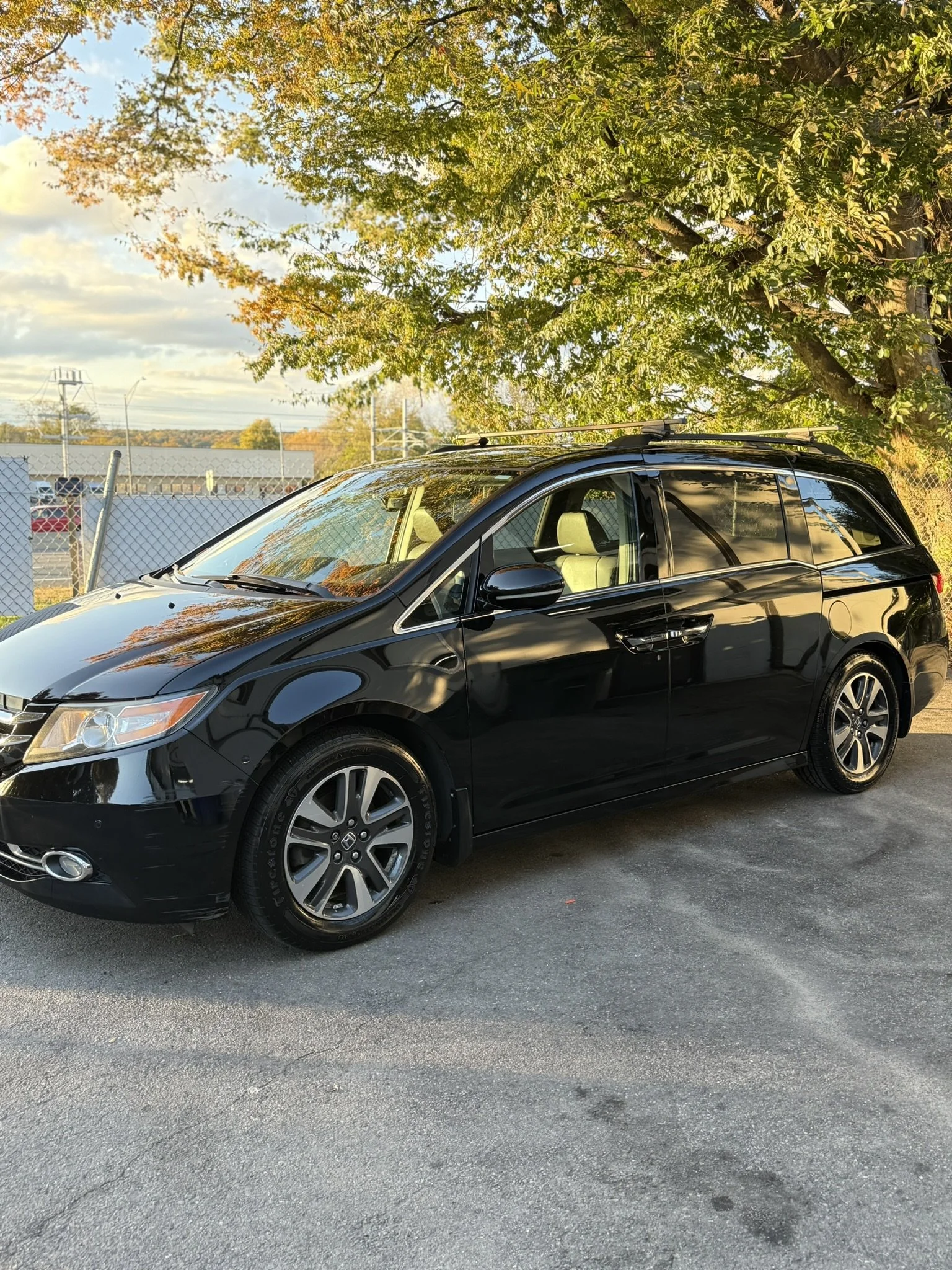 A black minivan parked on a paved lot near a tree with green and orange leaves under a partly cloudy sky.