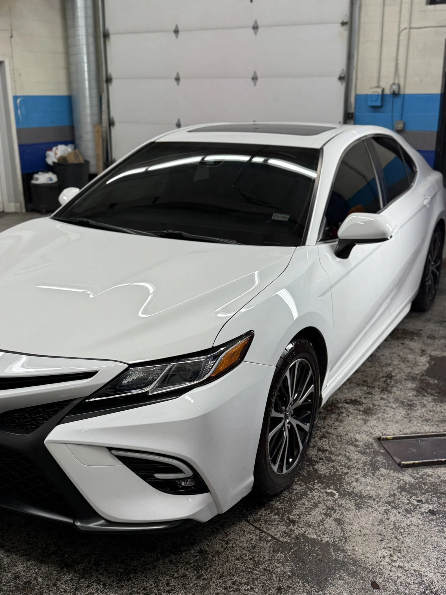 A white sedan parked inside a garage with a roll-up door, blue and gray painted walls, and various items including a trash can in the background.