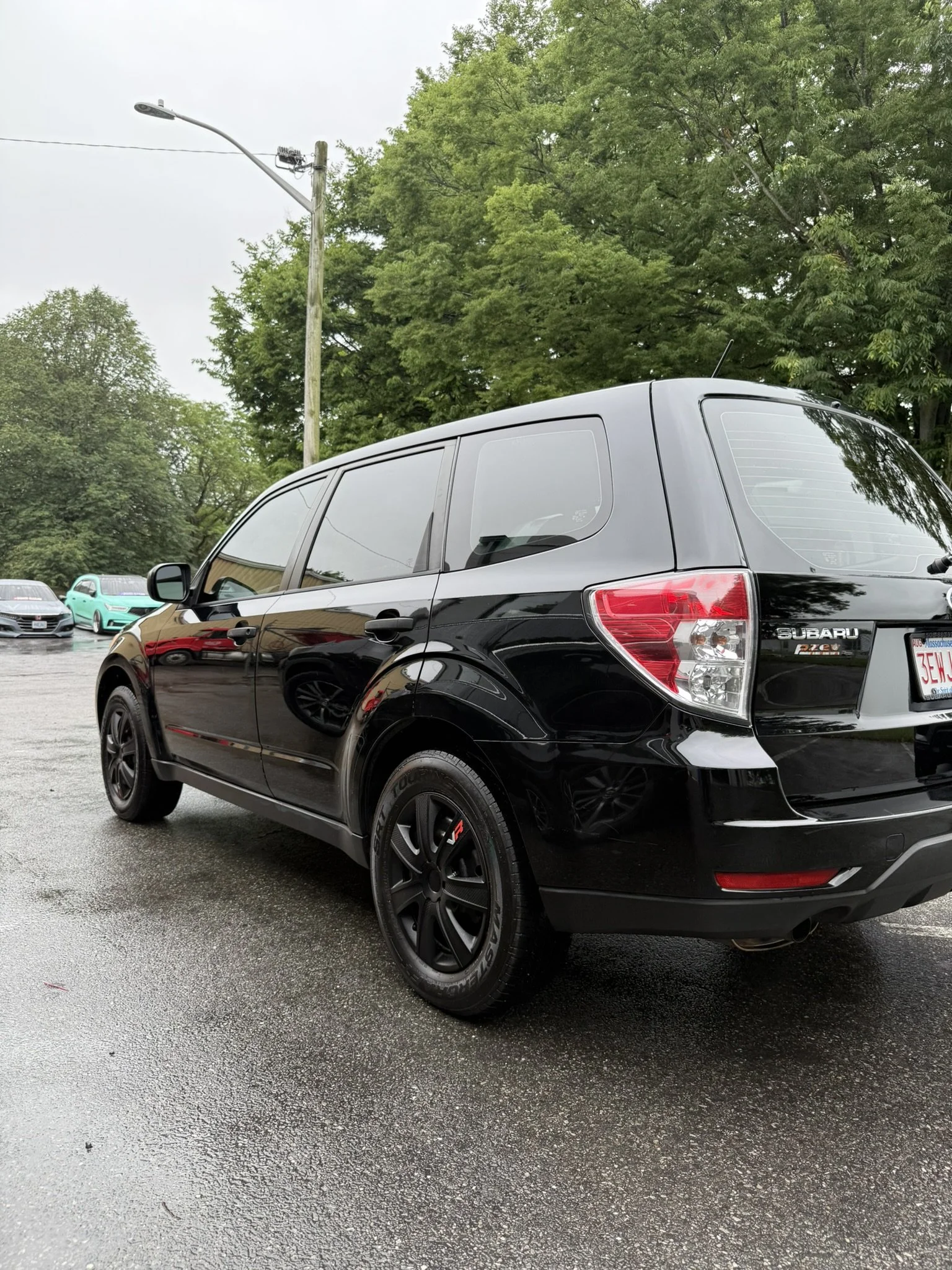 Black Subaru Outback parked on a wet asphalt lot with trees and other cars in the background.