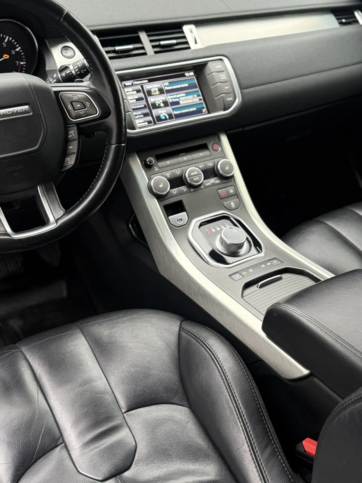 Interior of a modern vehicle showing a leather seat, dashboard, touchscreen display, and center console with gear selector and controls.