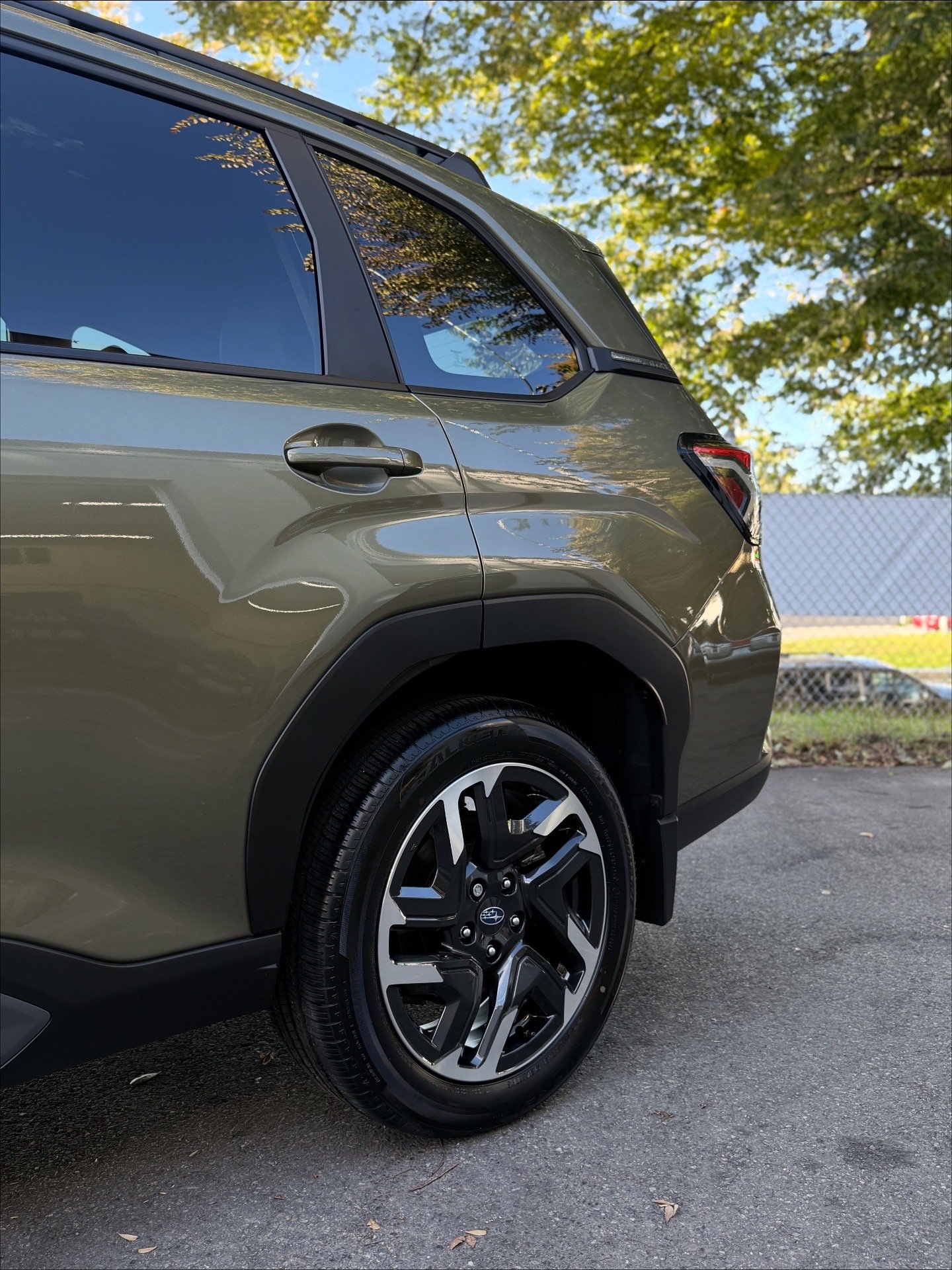 Close-up of the rear side of a green SUV, showing the wheel, tire, and part of the rear and side windows, with trees and a chain-link fence in the background.
