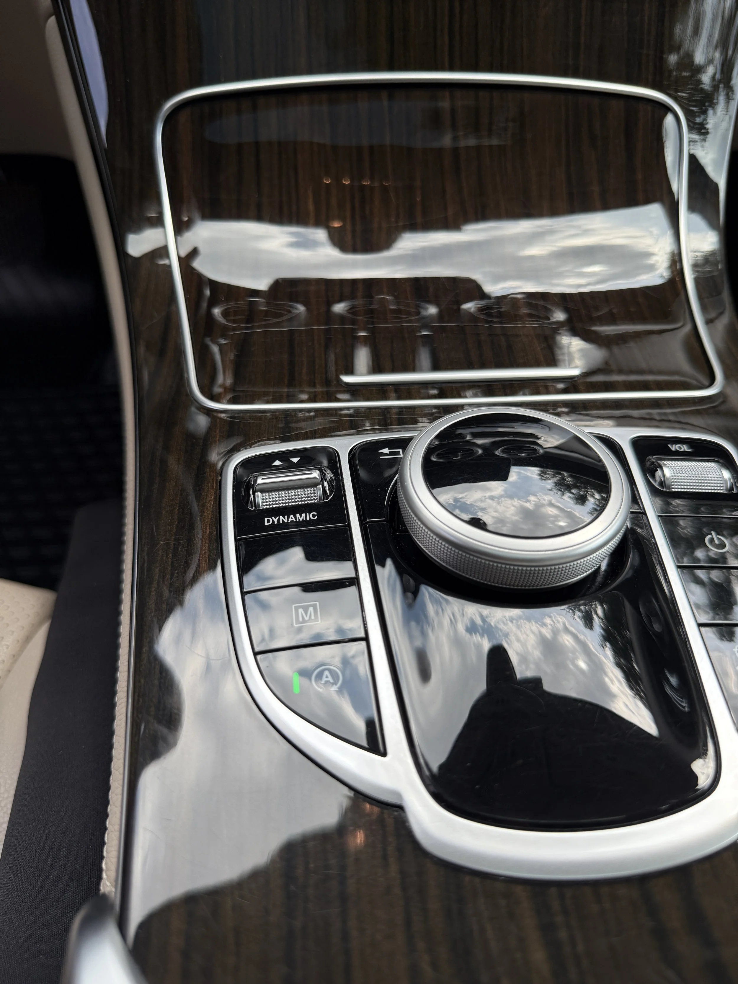 Close-up of a luxury car's center console with wood paneling, a large control dial, and various buttons, reflecting the sky and clouds.
