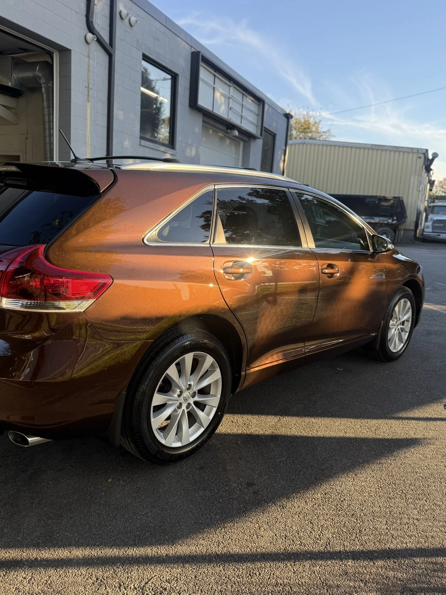 A brown station wagon parked outside a commercial building during sunset, with reflections of trees and sky on its surface.