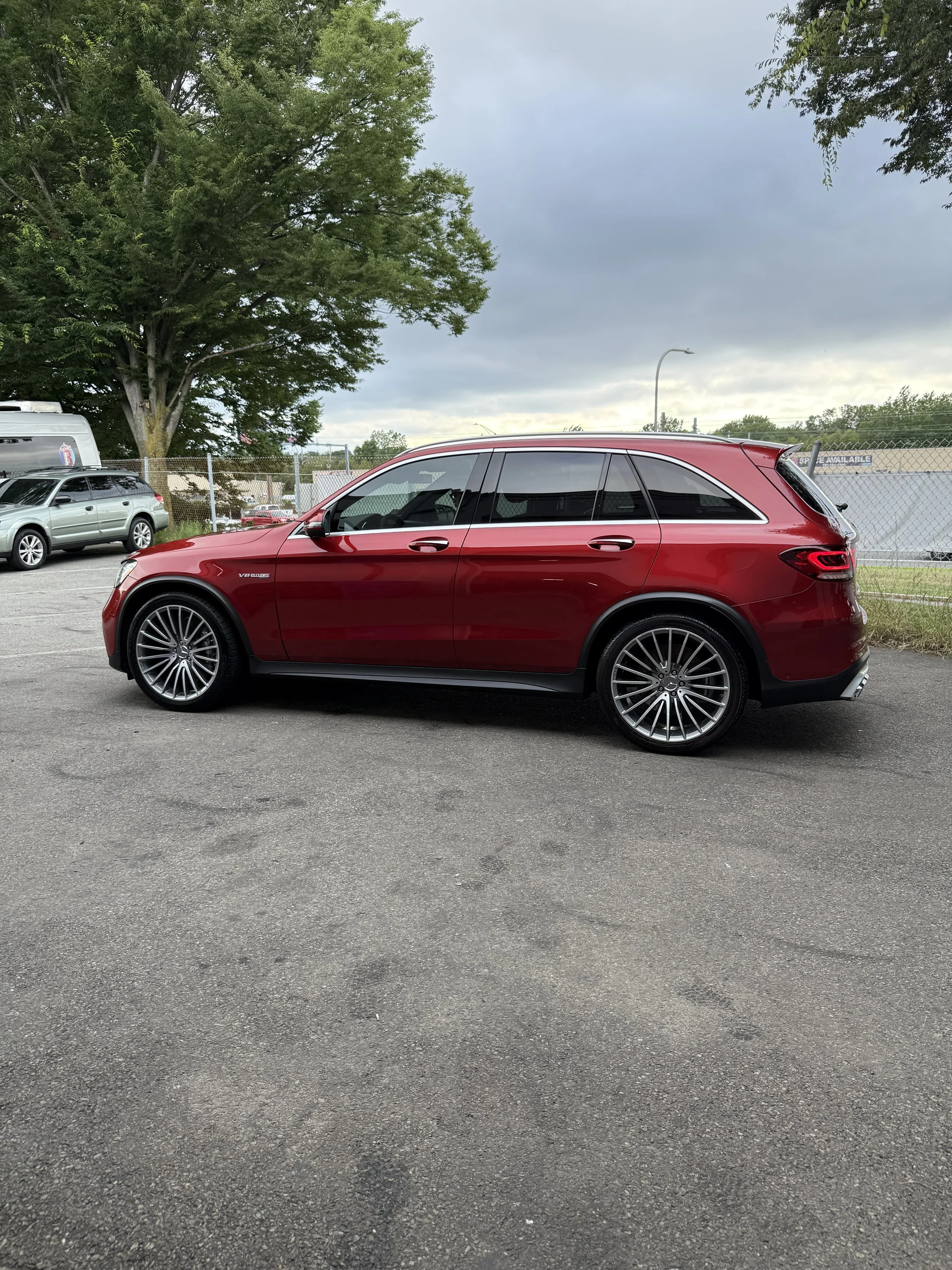 A red luxury SUV parked on a paved lot with trees, other cars, and a chain-link fence in the background under a cloudy sky.