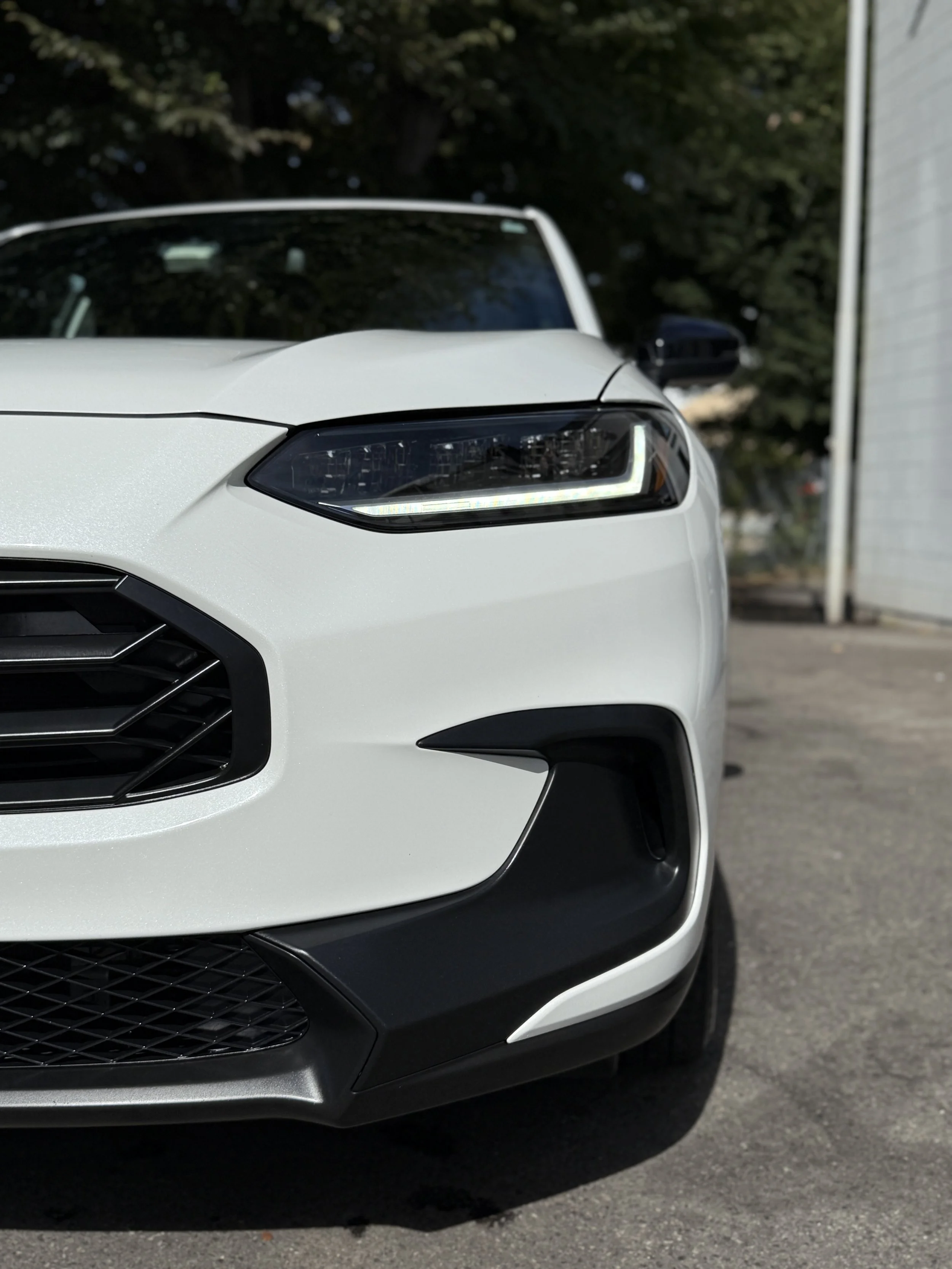 Close-up of the front left side of a white modern car showing its headlight, grille, and bumper in an outdoor setting.