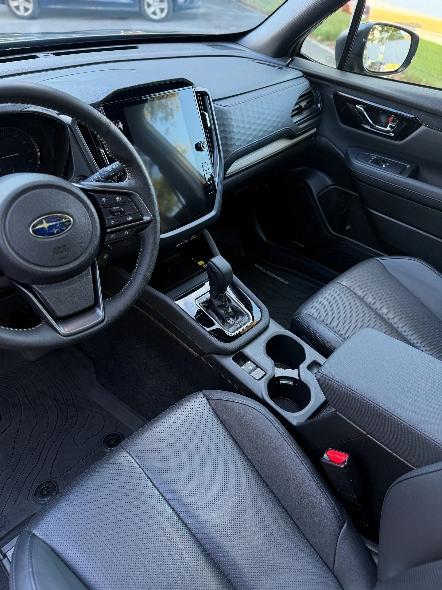 Interior of a modern Subaru vehicle showing the driver's seat, steering wheel, central console with gear shift, large touchscreen display, and cup holders, with outdoor scenery visible through the windshield and side window.