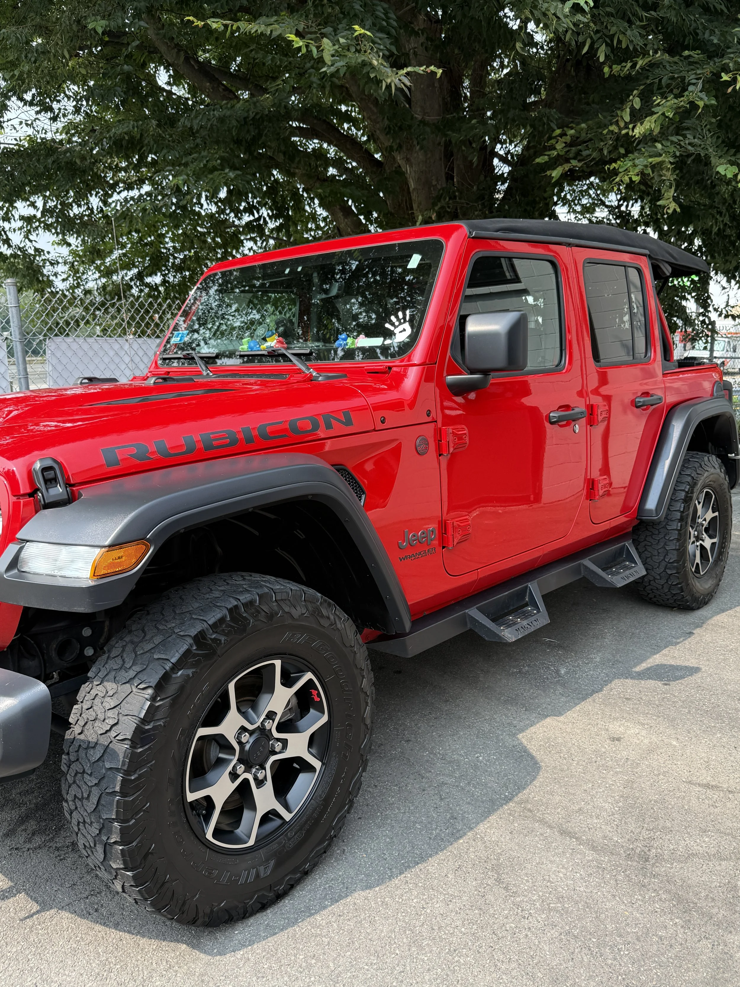 Red Jeep Rubicon with black fender flares, four doors, black Matt tire with black and silver rims, parked on street near green trees.