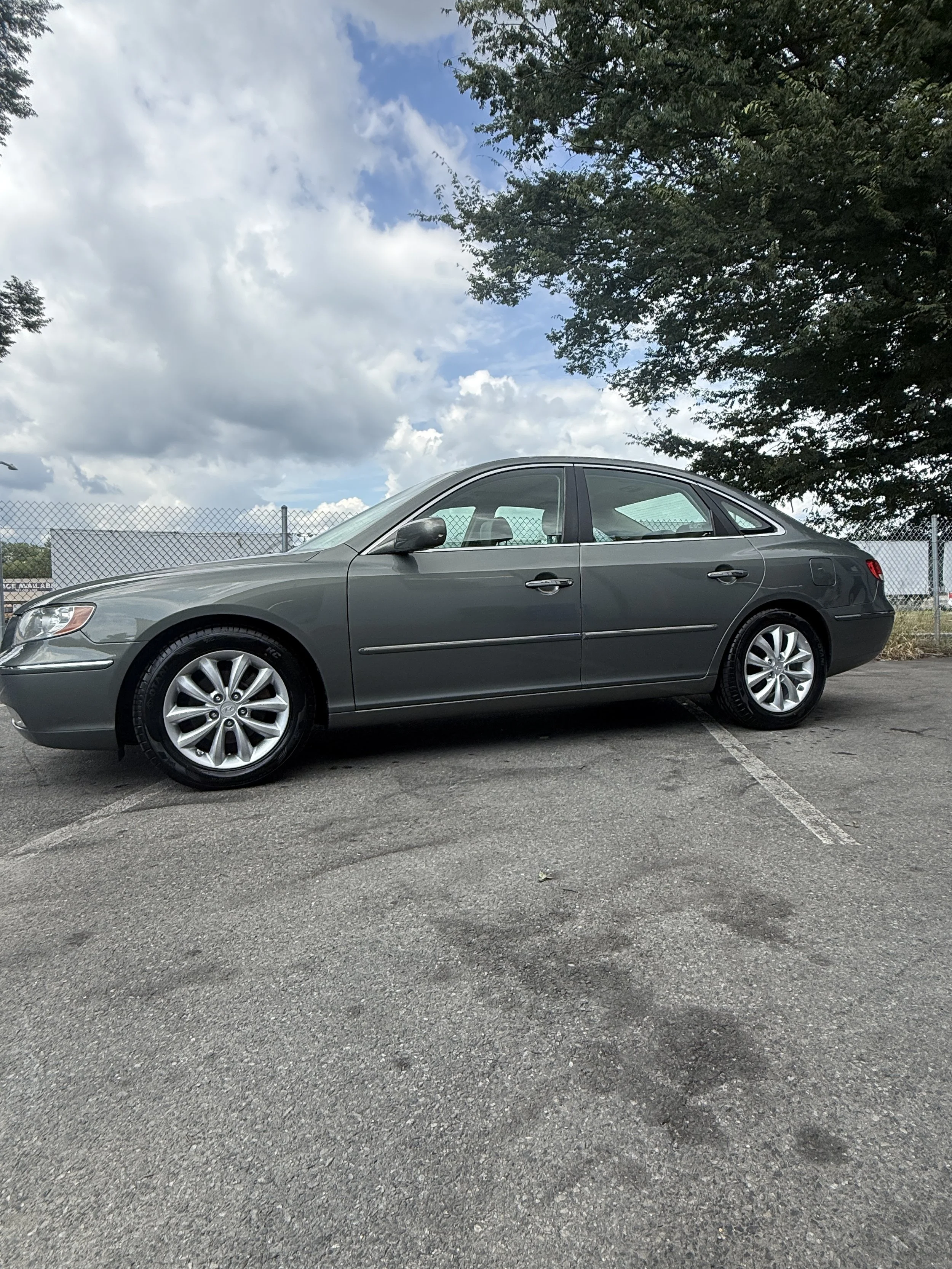 A dark gray, four-door sedan parked in a parking lot under a partly cloudy sky with trees and a chain-link fence in the background.