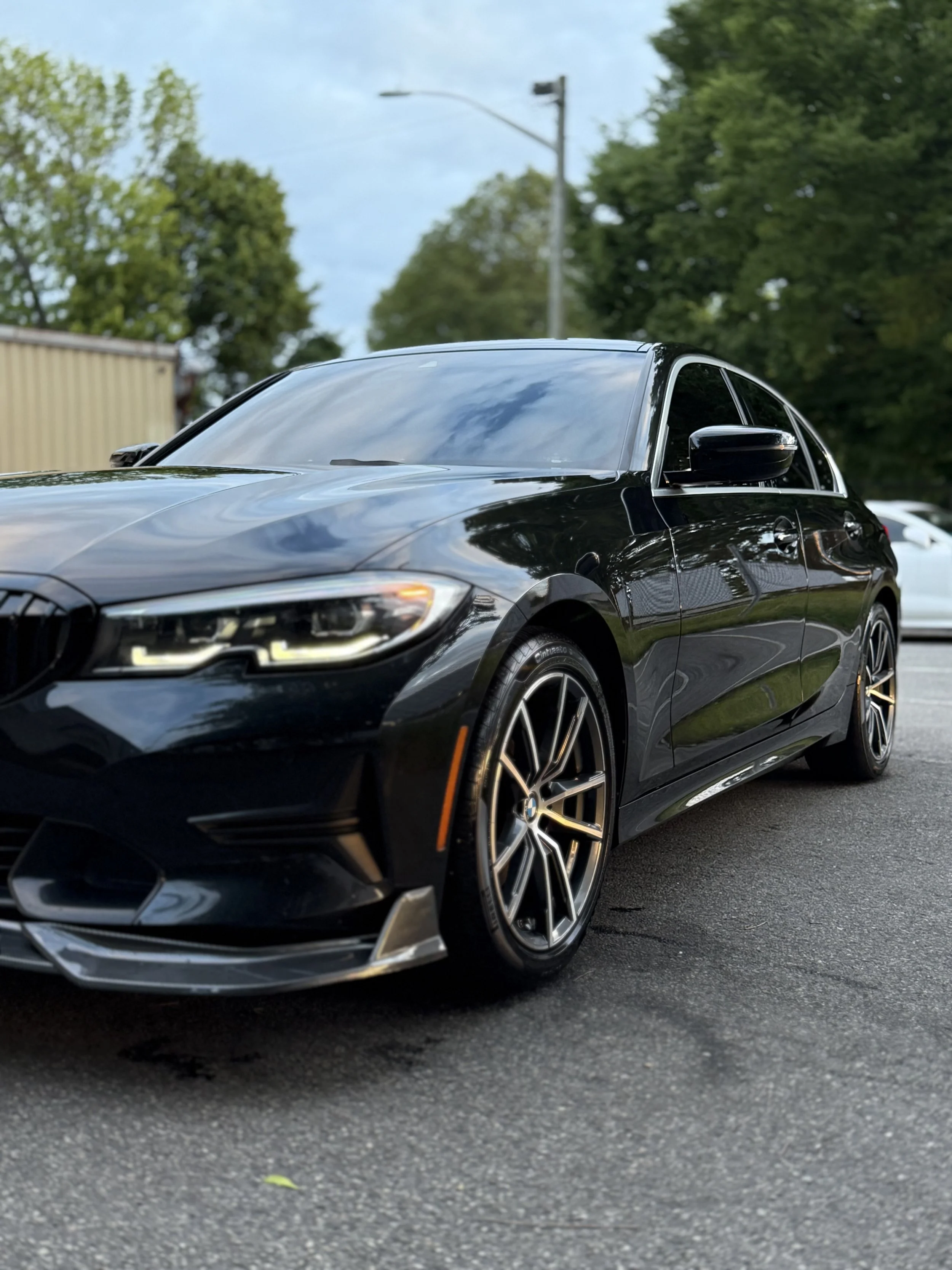 Black modern luxury sedan parked on asphalt at an outdoor location during daytime, with trees and a cloudy sky in the background.