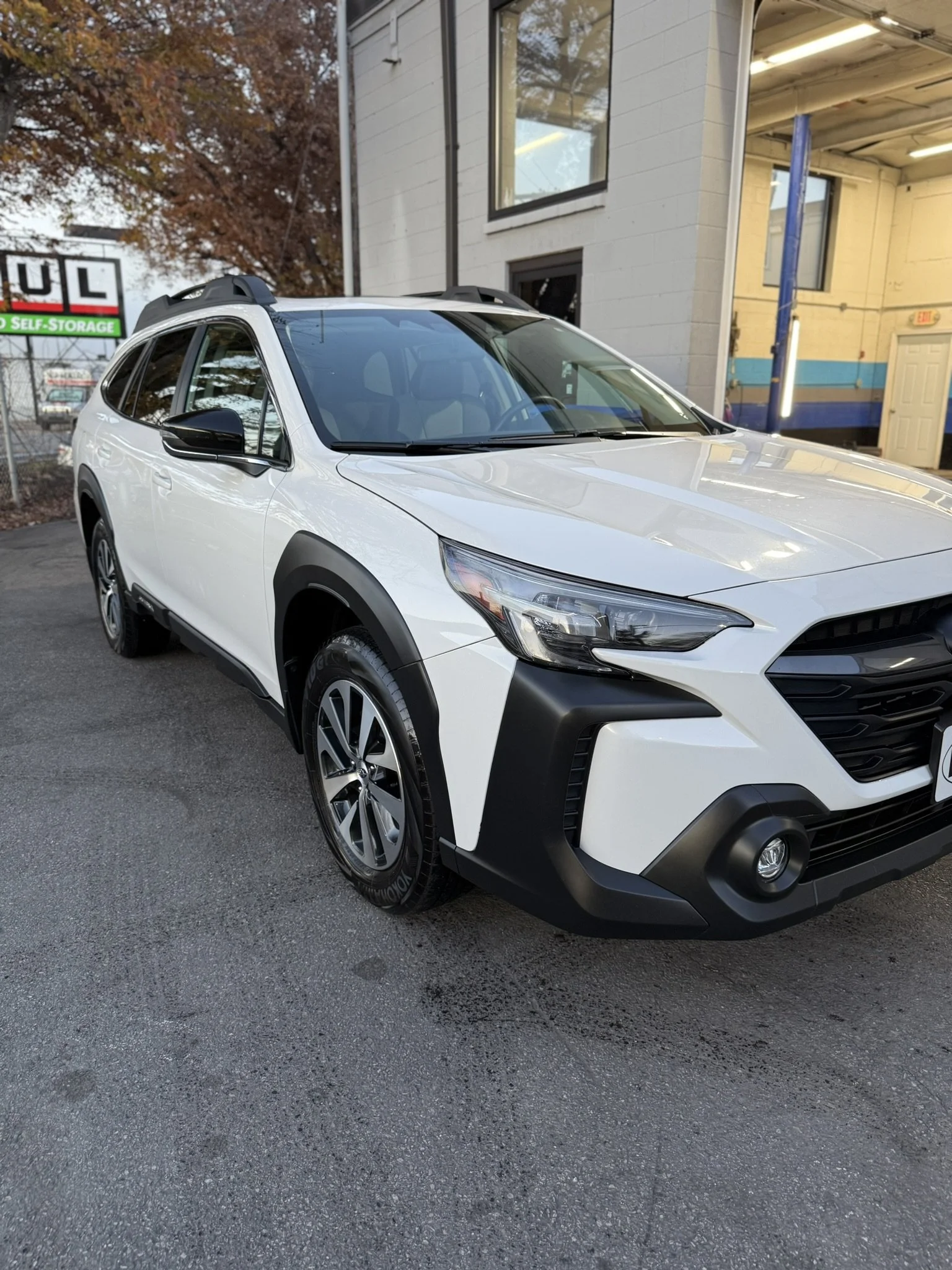 A white Honda station wagon parked on a paved lot near a building with a sign behind it that reads "U-HAUL Self-Storage". The building has a white and beige exterior with a blue stripe, and there are trees with autumn leaves in the background.