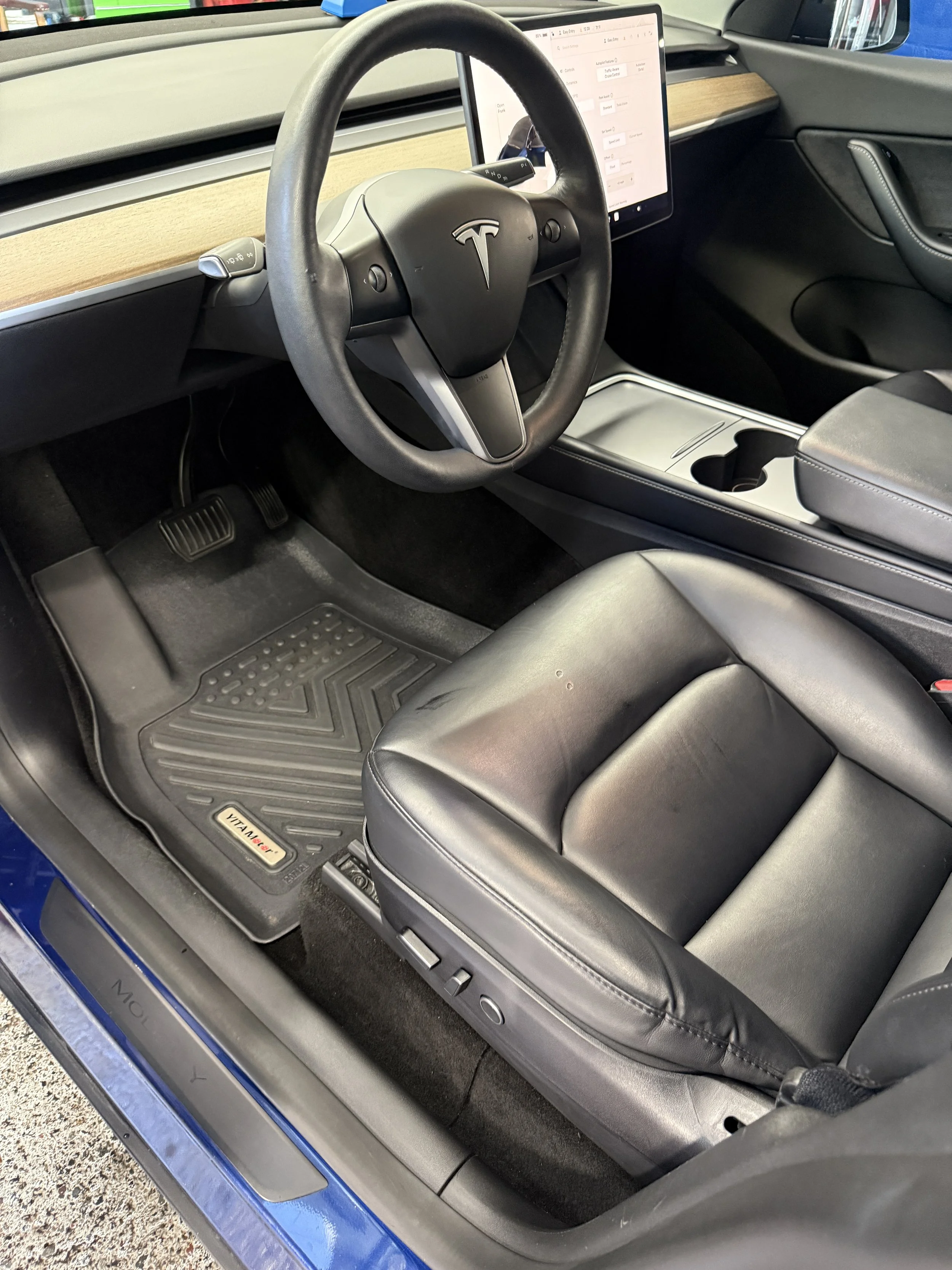 Inside view of a Tesla Model Y vehicle showing the steering wheel, dashboard, touchscreen display, and front seats with a black floor mat.