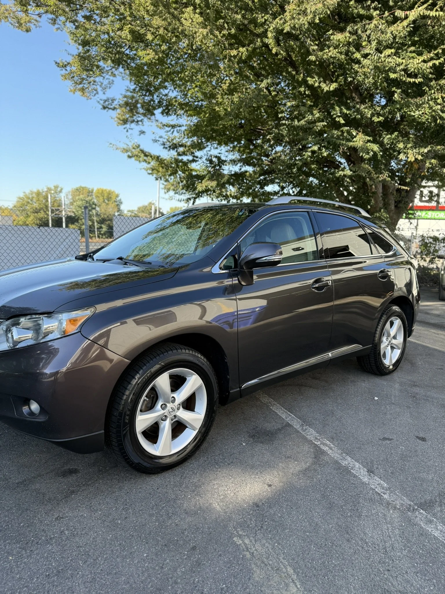 A black Mazda hatchback car parked in a parking lot on a sunny day, with a large green tree and clear blue sky in the background.