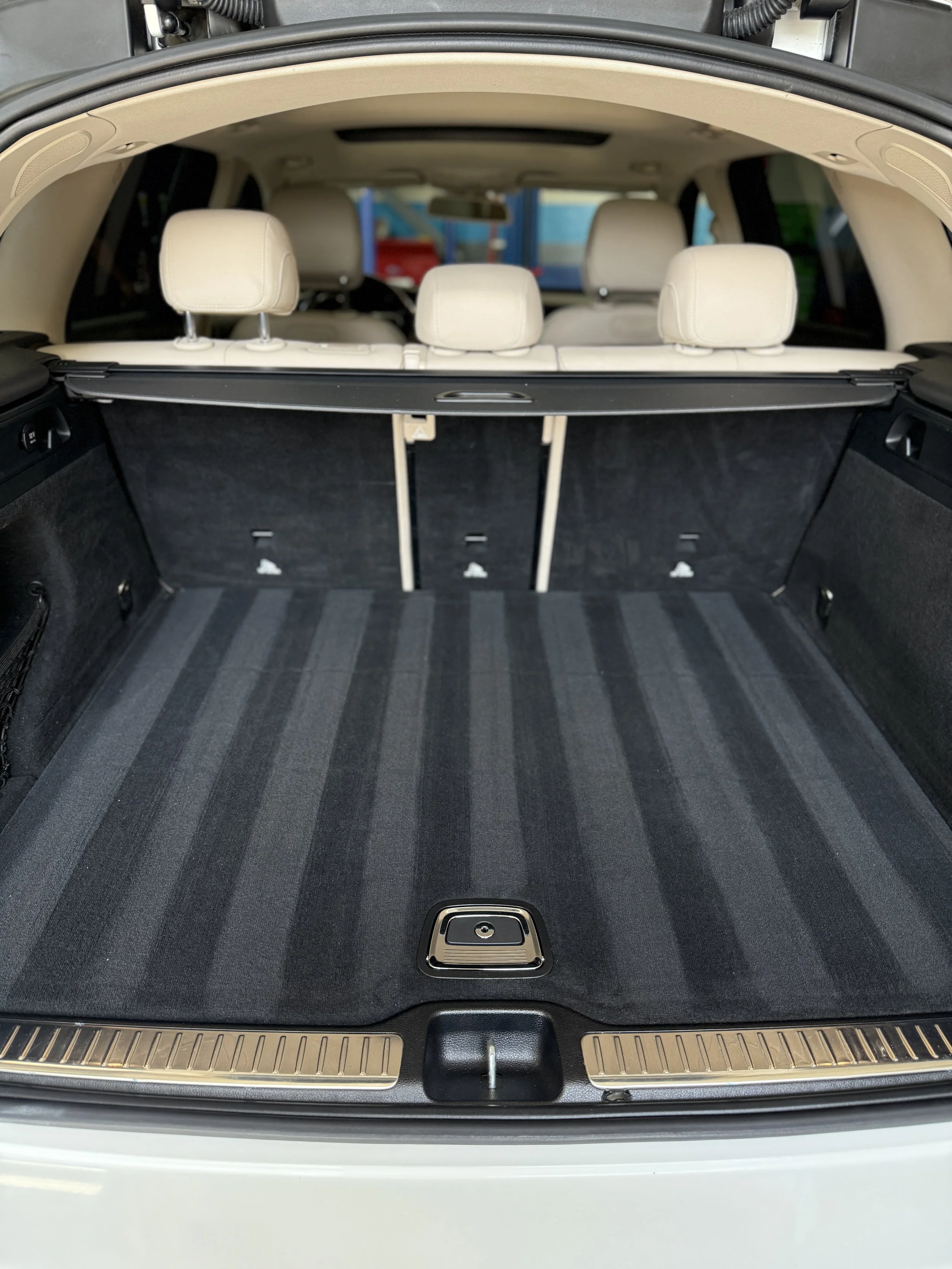 The photo displays the cargo area of a luxury SUV with a clean, striped, black and gray carpeted trunk. The seats are beige, and a black retractable cover is in place behind the rear seats.