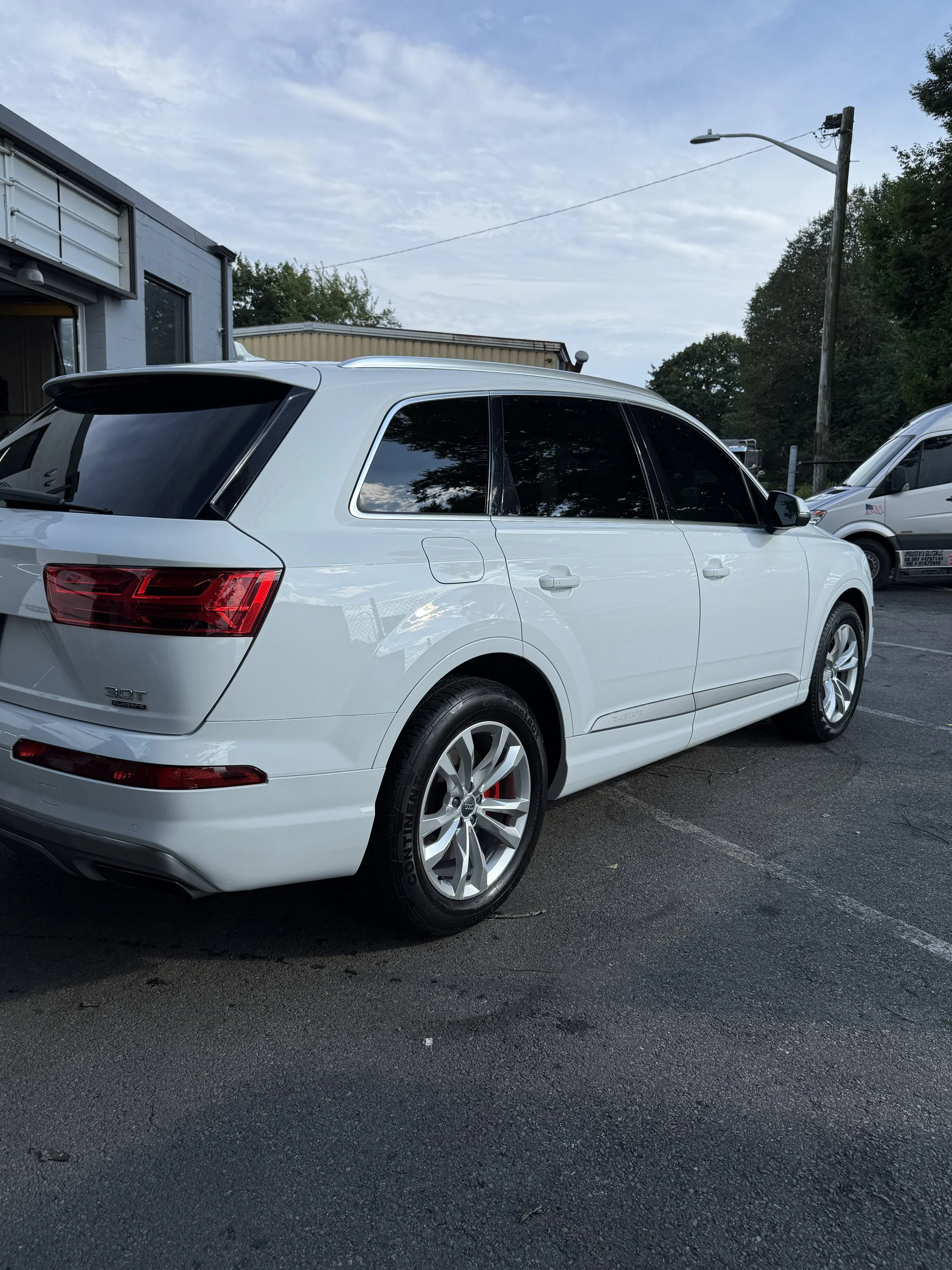 White SUV parked in a parking lot with a building and trees in the background, under a partly cloudy sky.