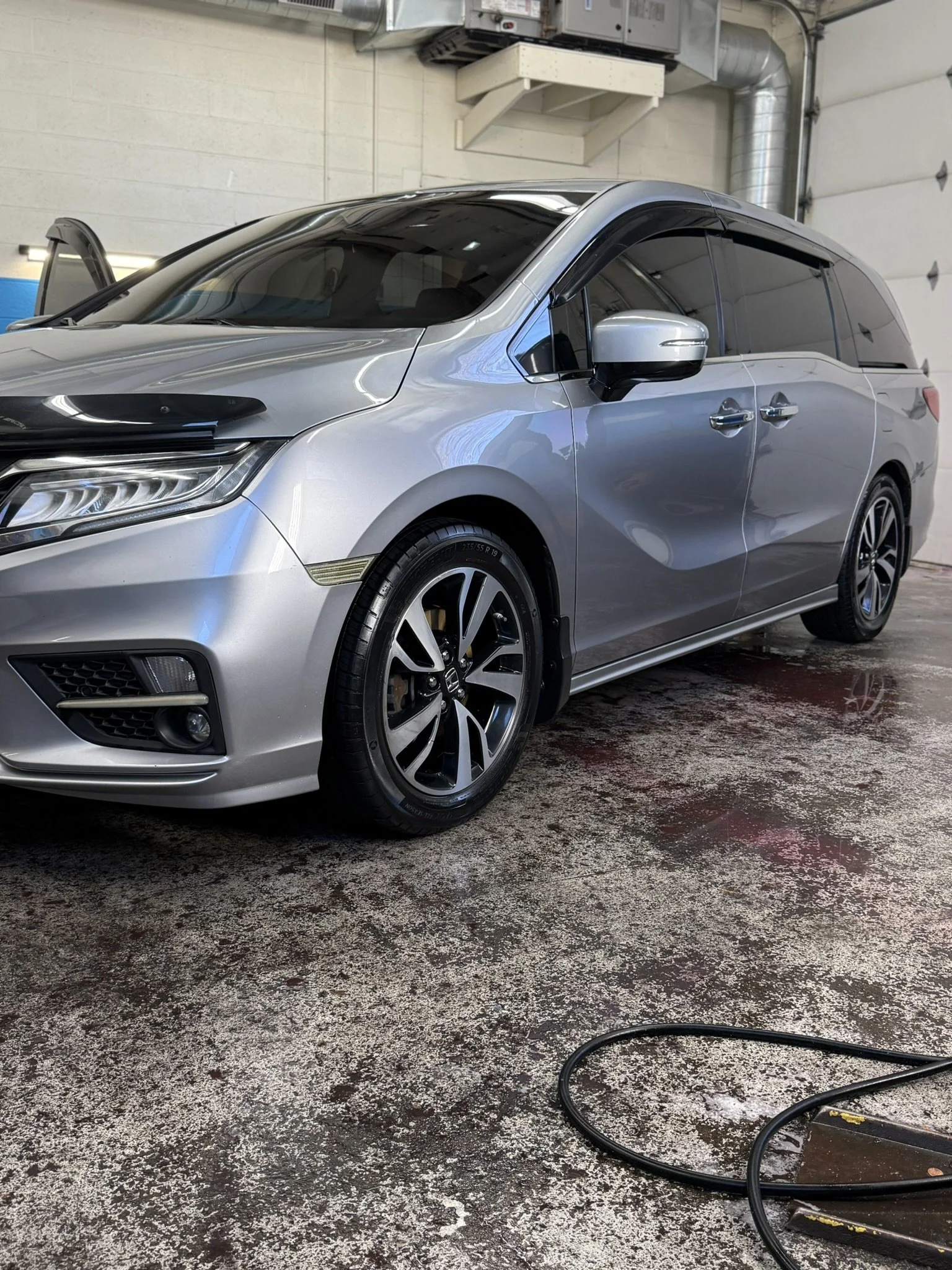 A silver Honda minivan inside a garage or car wash with wet floor and a black water hose on the ground.