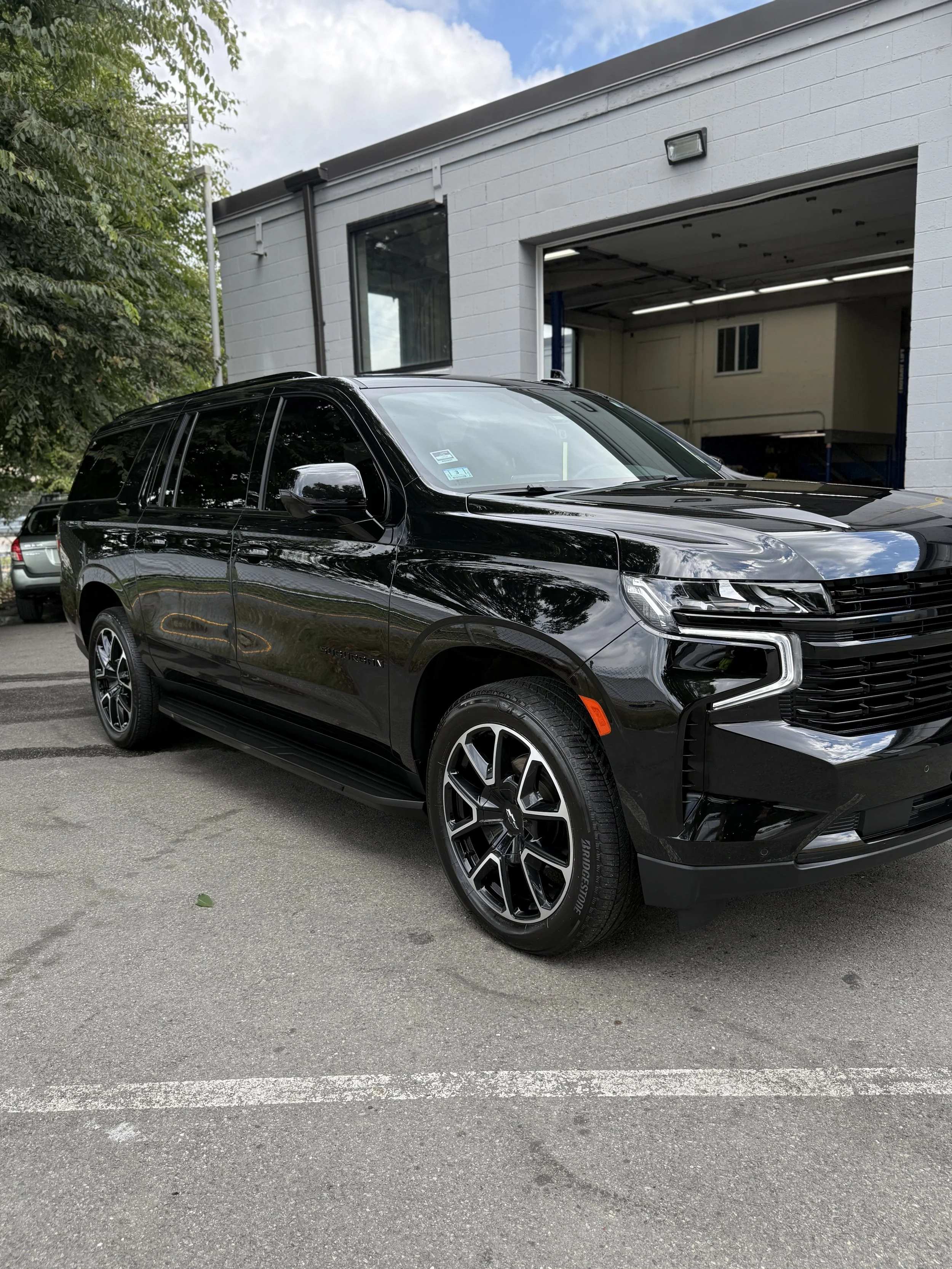 A black Chevrolet Suburban parked outside a white garage with an open door in a parking lot.