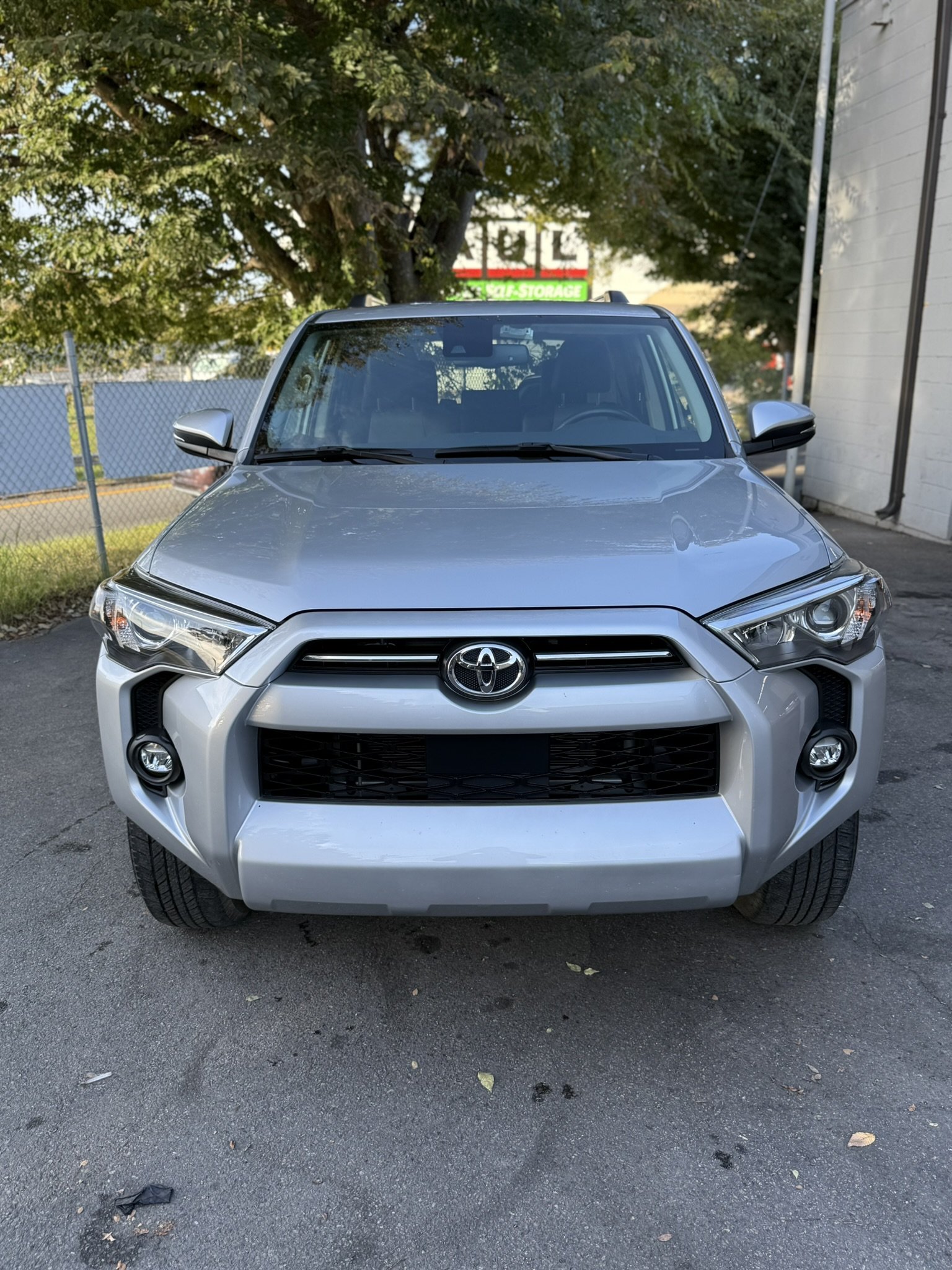 Front view of a silver Toyota SUV parked on asphalt near a white building with a tree and a chain-link fence in the background.