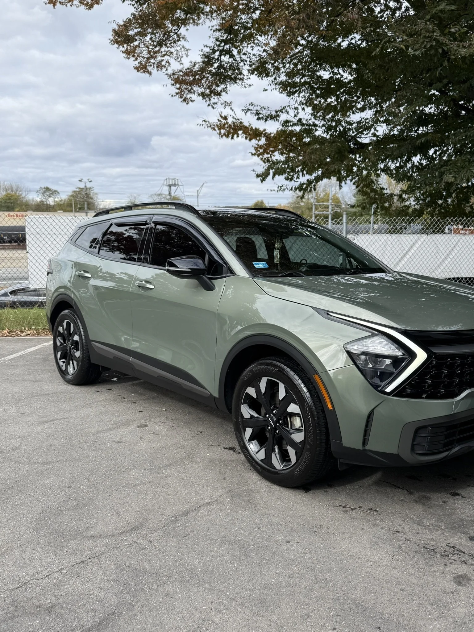 A modern green SUV parked on a paved surface under a tree with a chain-link fence and objects in the background.