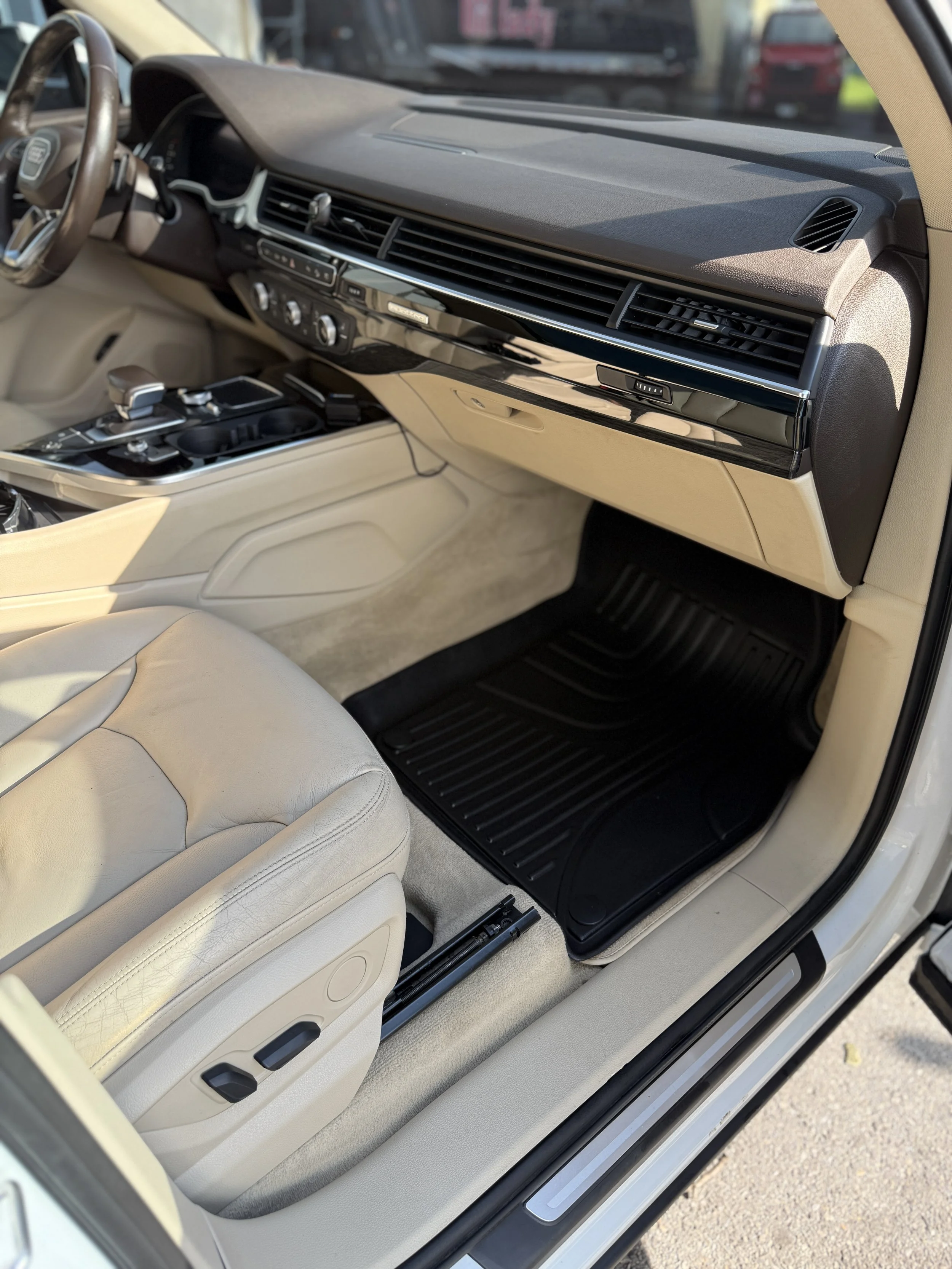Interior of a luxury car with beige leather seats, black dashboard, and black floor mats, showing the passenger side with the seat adjustment controls visible.