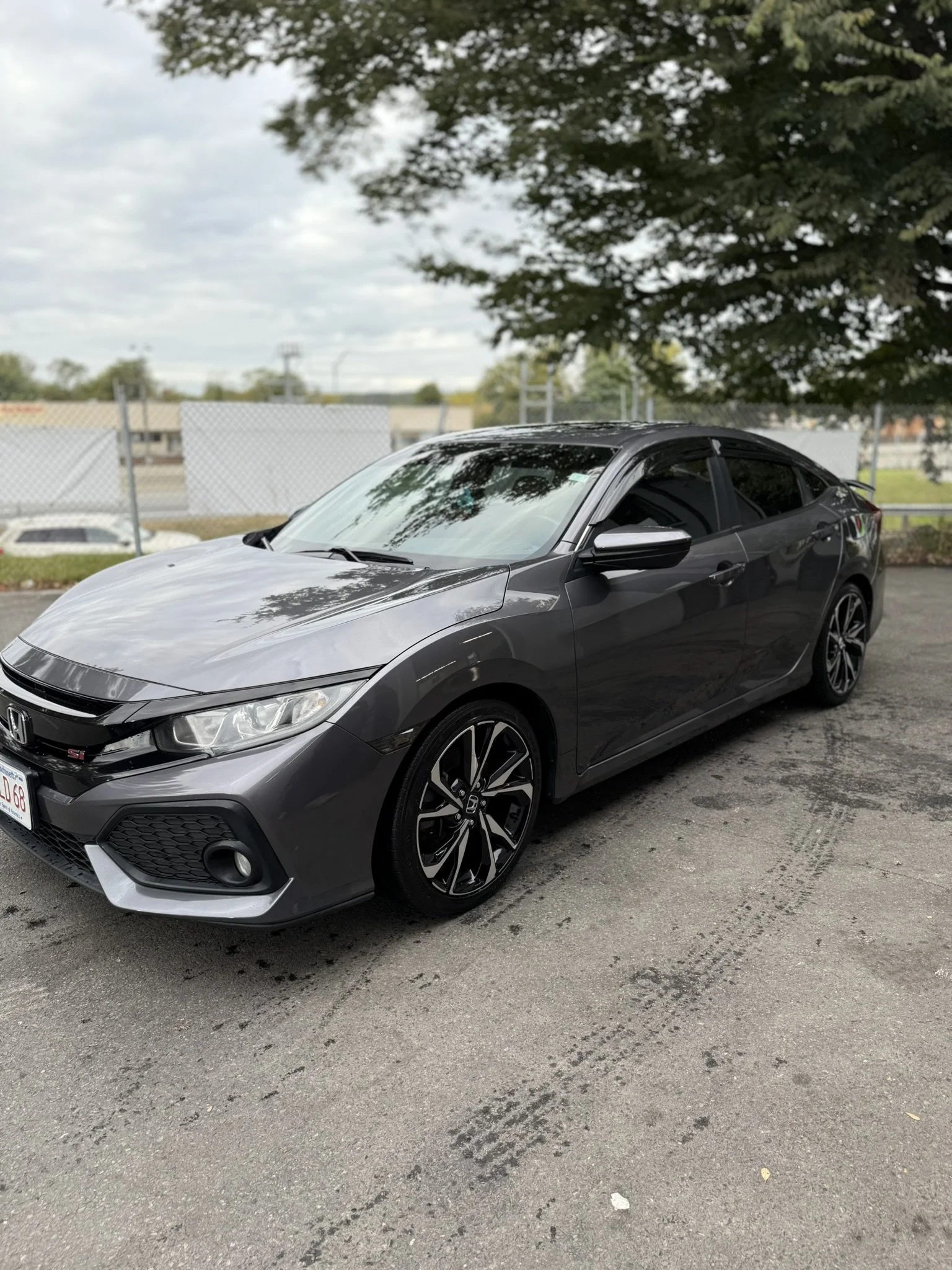 A dark gray Honda Civic sedan parked on a paved lot under a tree with overcast sky in the background.