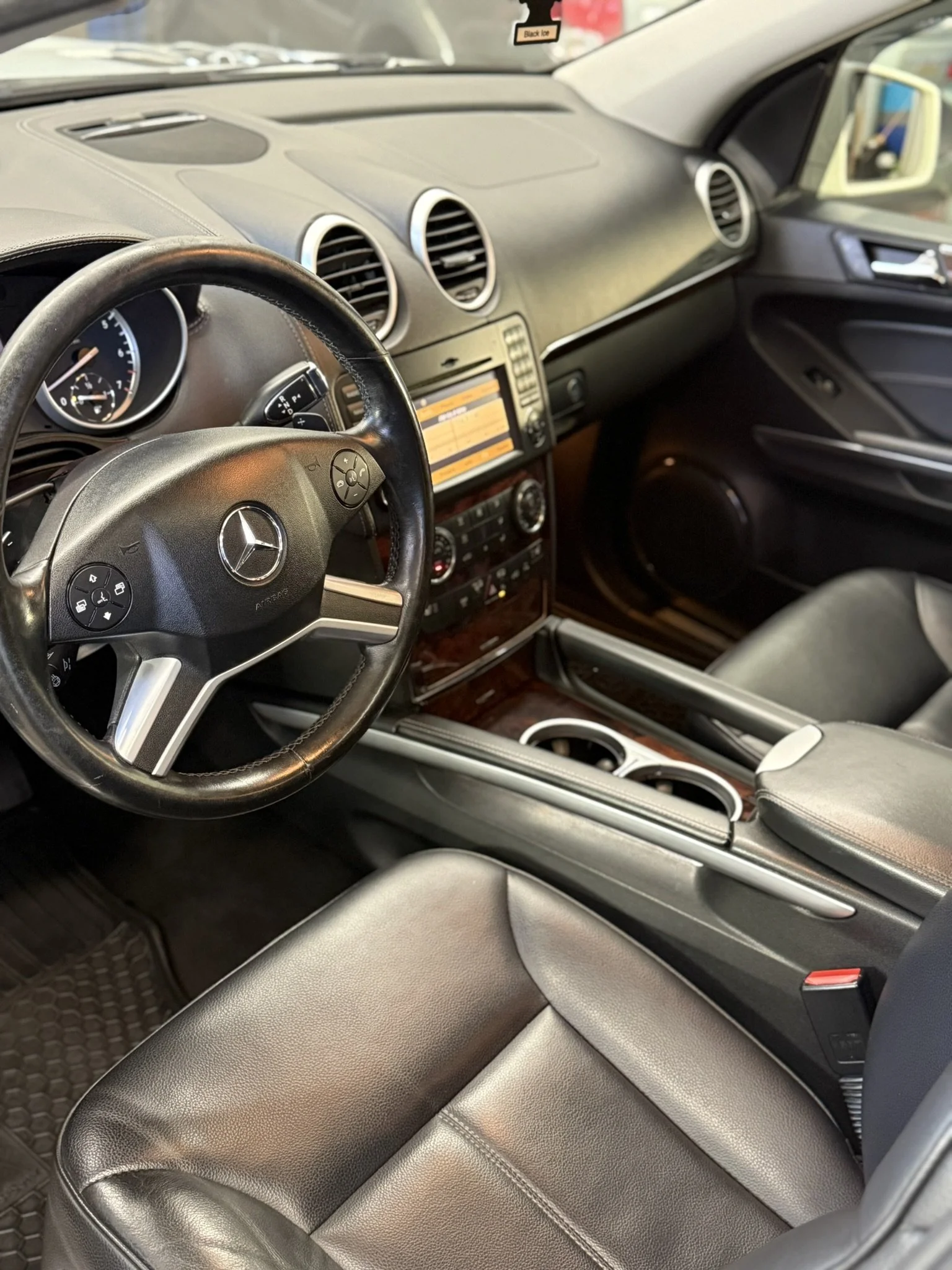 Interior of a Mercedes-Benz vehicle with black leather seats, a steering wheel with the Mercedes logo, and a dashboard with a navigation screen, air vents, and control buttons.