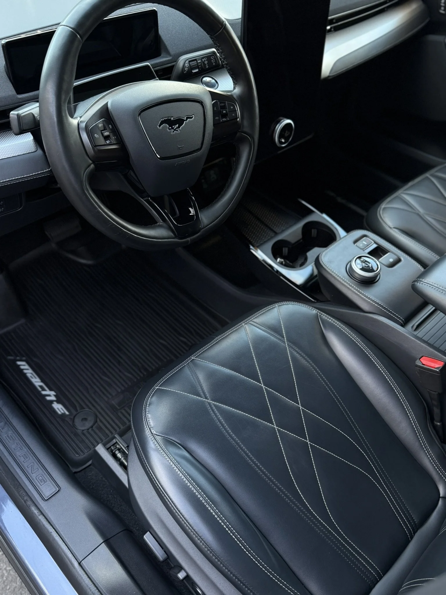 Interior of a Ford Mustang Mach-E electric vehicle, showing the steering wheel with Mustang logo, dashboard, gear selector, center console with cup holders, and the front seat with diamond-stitched black leather upholstery.