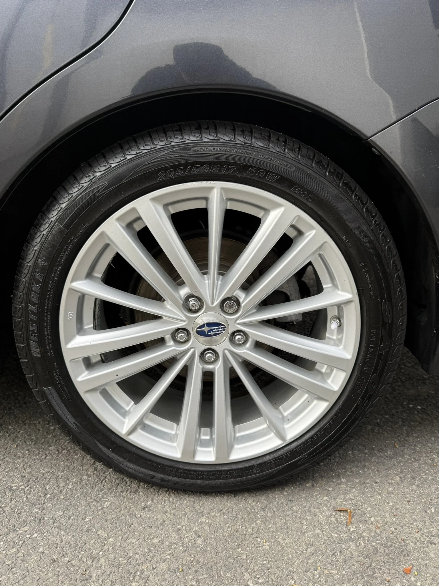 Close-up of a car tire and silver alloy wheel with the Subaru logo, parked on a gray asphalt surface.