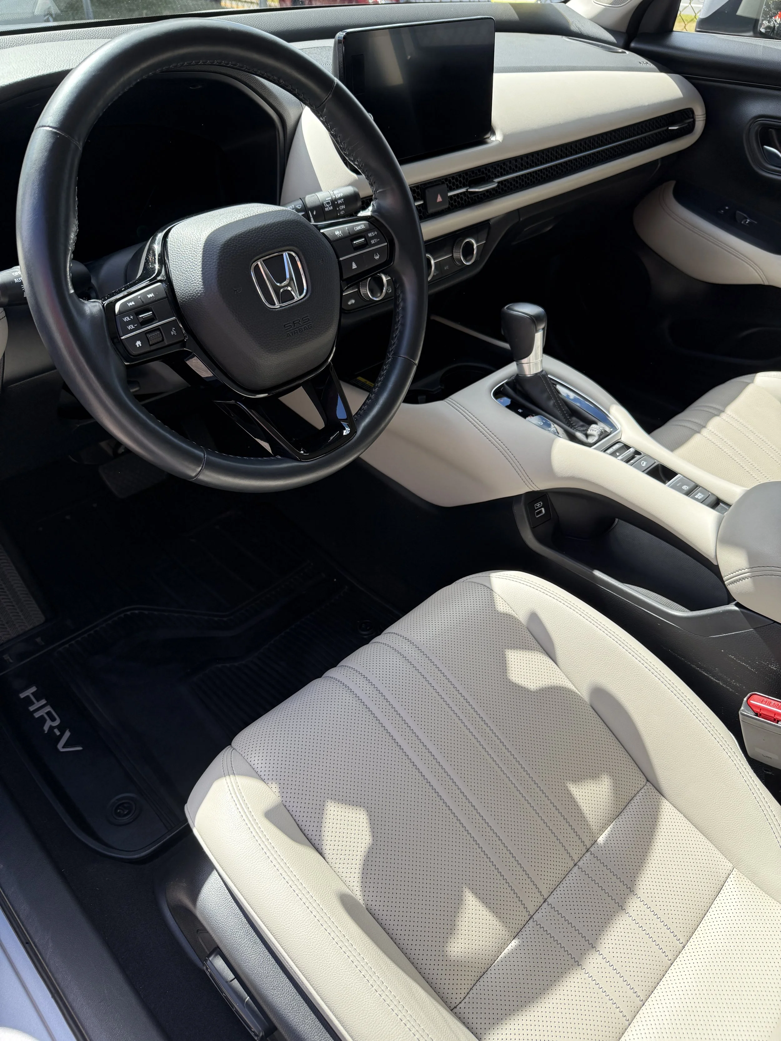 Interior of a Honda HR-V featuring a black steering wheel with the Honda logo, a digital display screen on the dashboard, a gear shift, and beige leather seats with perforated upholstery.