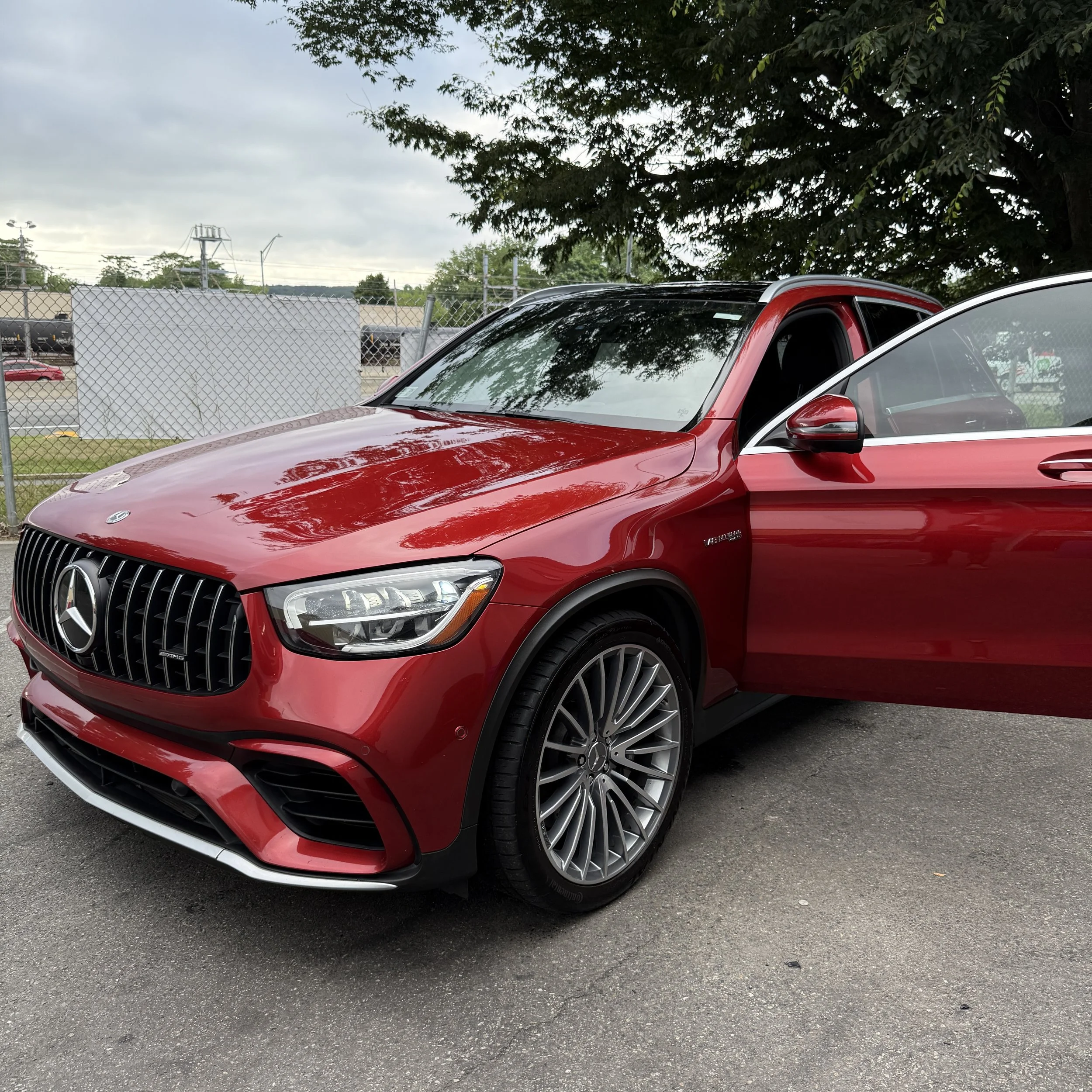 Red Mercedes-Benz SUV parked outdoors with one front door open, set against a backdrop of trees, a chain link fence, and overcast sky.