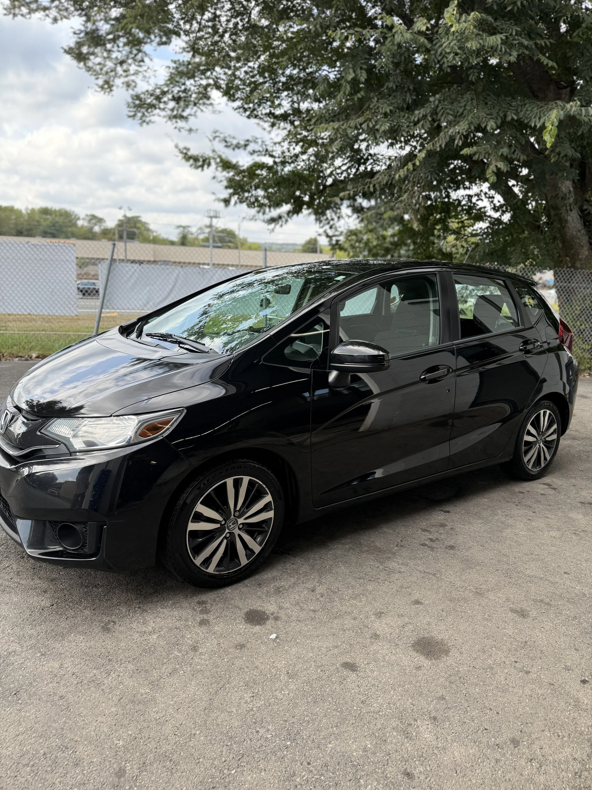 Black Honda hatchback car parked outdoors on a paved surface with trees and a chain-link fence in the background.
