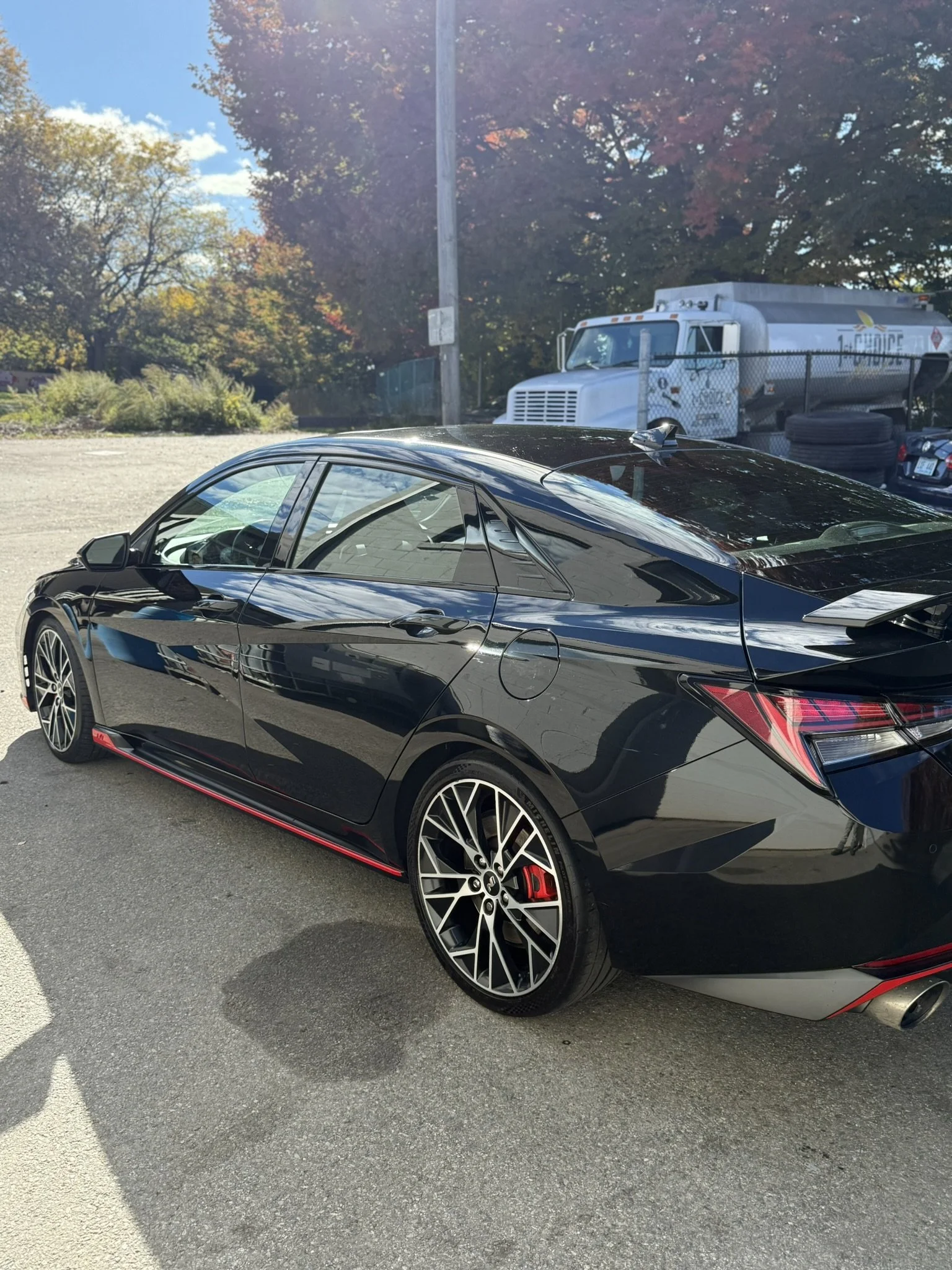 A black sports car with red accents parked outdoors on a sunny day, with trees displaying fall foliage in the background.