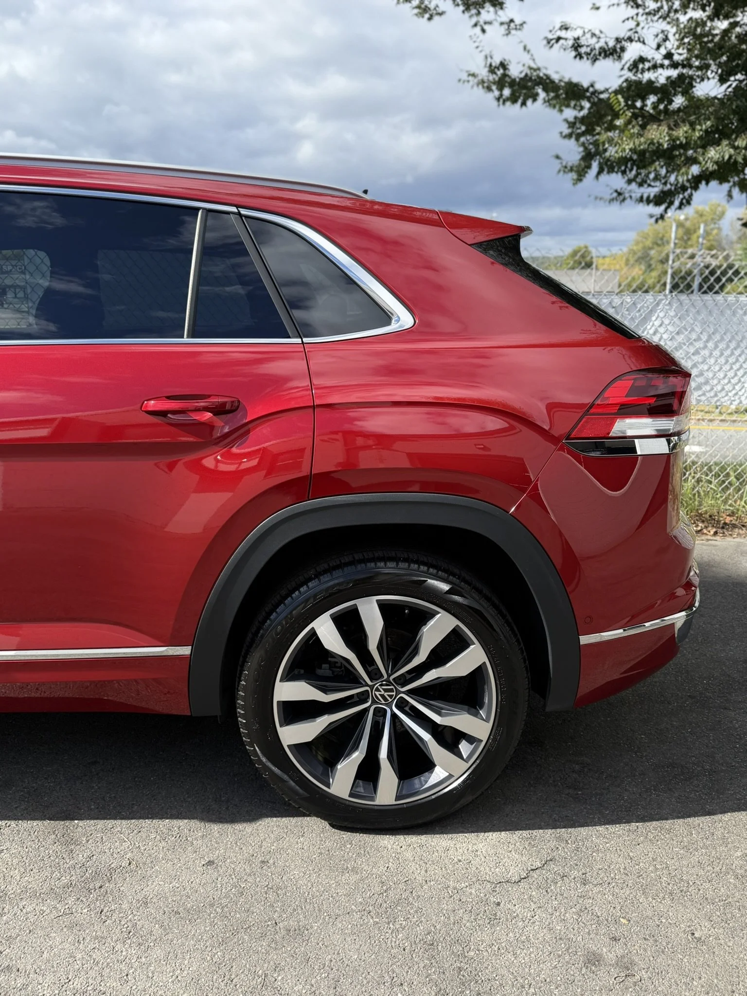Close-up of the rear side of a red SUV with black and silver alloy wheels, parked on a paved surface next to a chain-link fence, with a partly cloudy sky in the background.