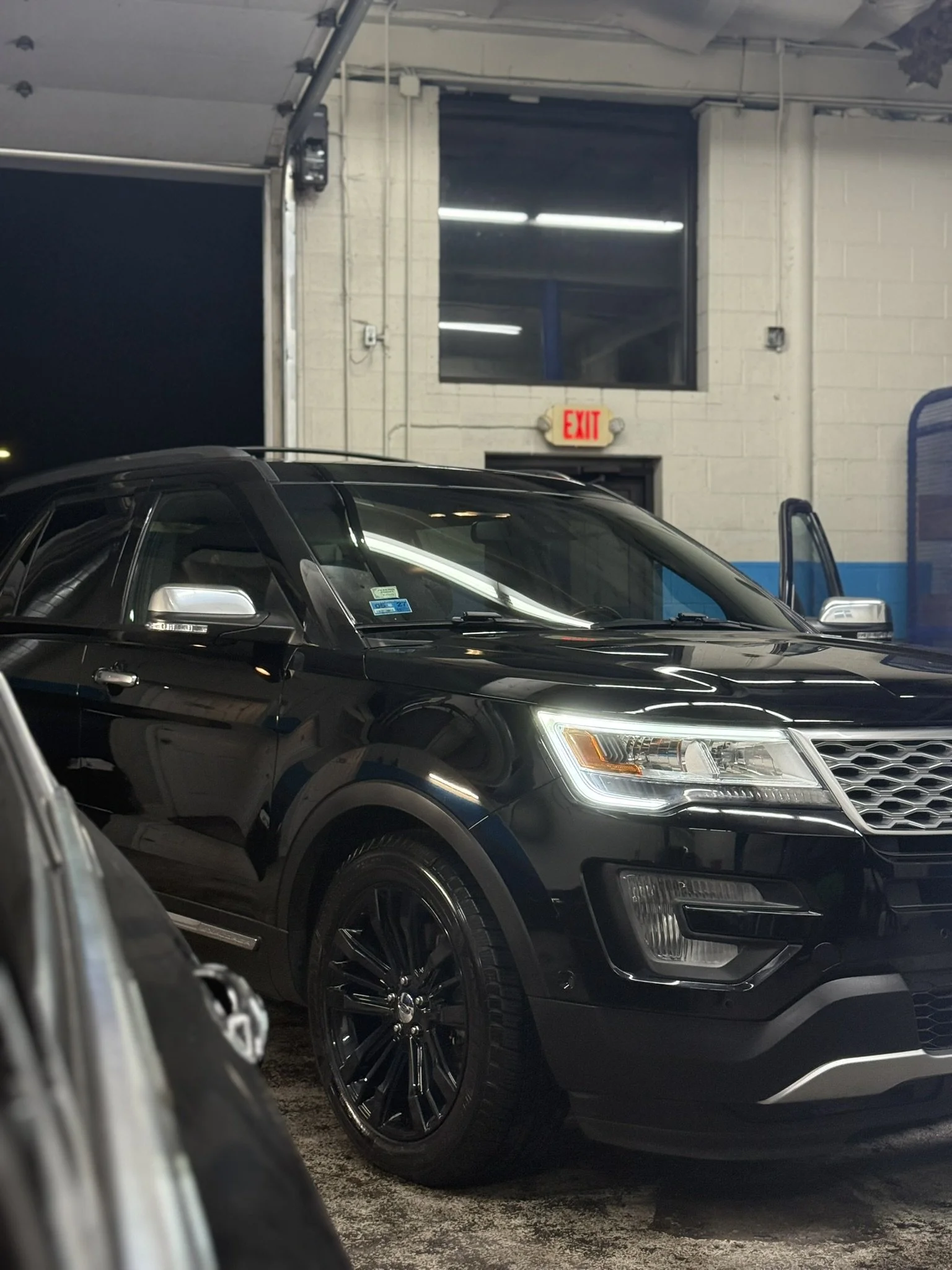 Black SUV parked inside a garage with a white brick wall, a window, an illuminated exit sign, and fluorescent ceiling lights.
