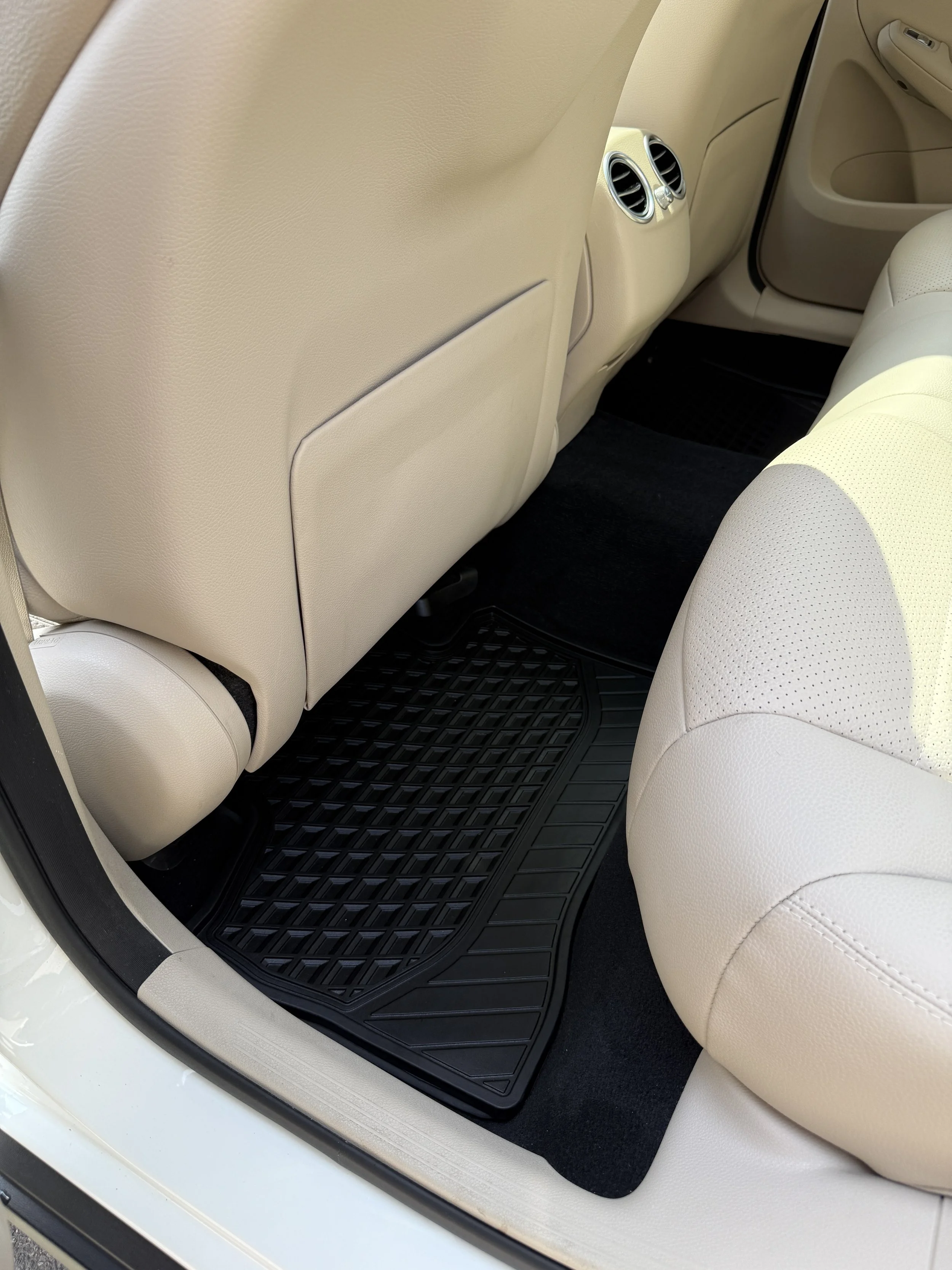 The back seat area of a beige leather car interior with black floor mats and front seat backrests.