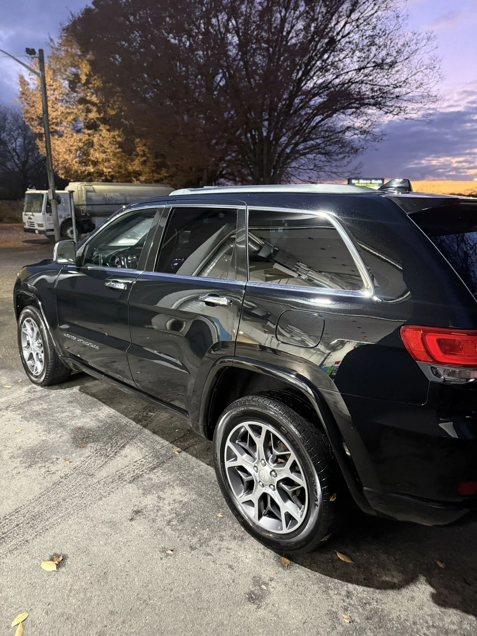 A black SUV parked on a dirt lot during dusk with a large tree and a water tanker truck in the background.