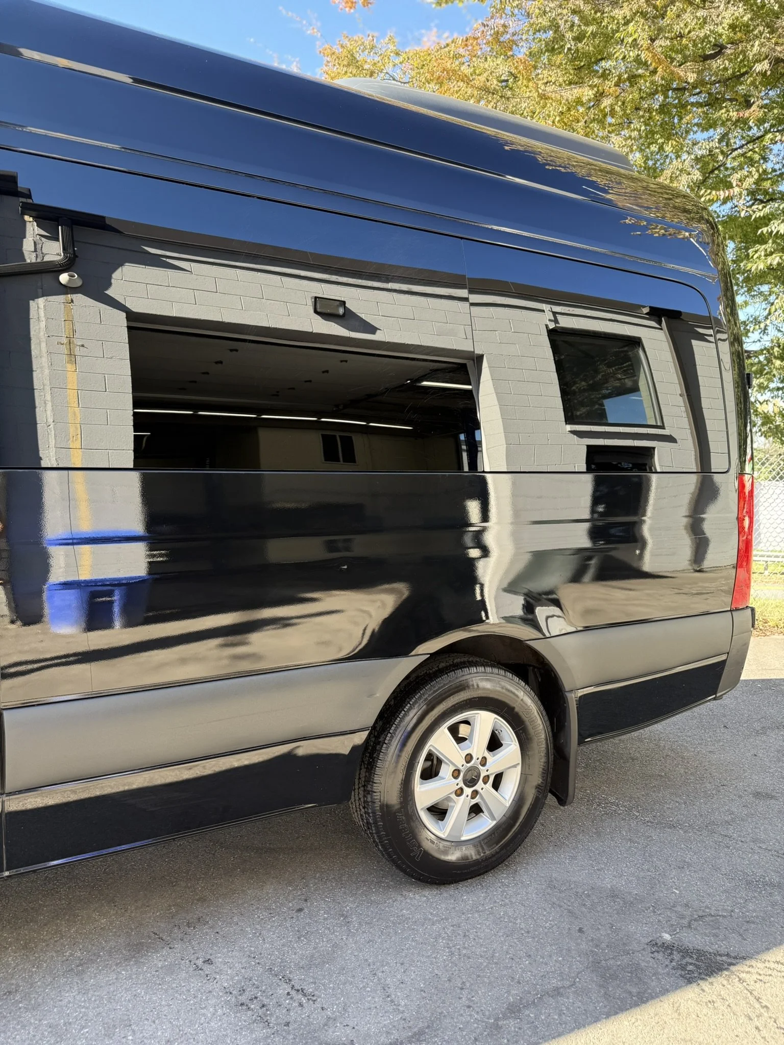 A black van with a partially open side window shows an indoor garage area with a gray brick wall and a small window, reflected on the van's shiny surface. There are trees with green and yellow leaves in the background and a clear blue sky.