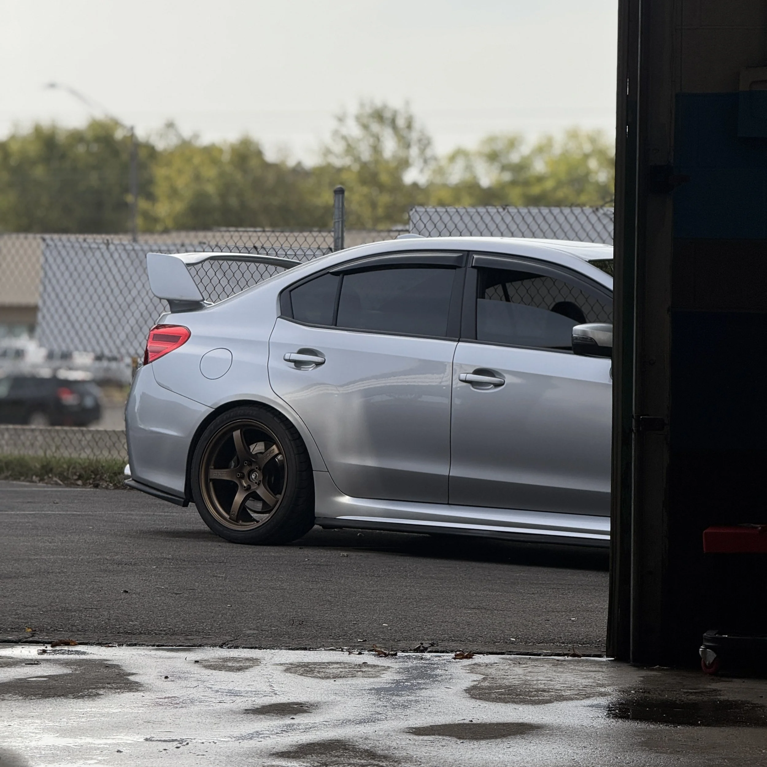 Silver sports car with a large rear spoiler, parked near a chain-link fence and partially inside a garage, with dark tinted windows and bronze-colored wheels.