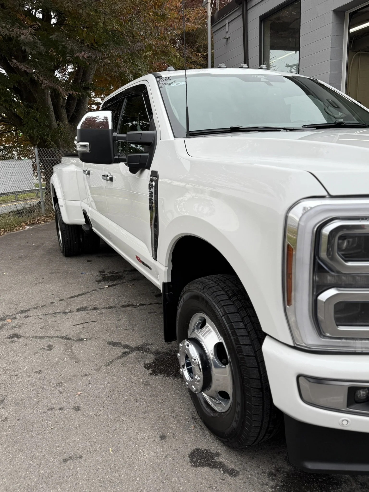 Front and side view of a white pickup truck parked on an asphalt lot with a gray building, a chain-link fence, and trees with fall foliage in the background.