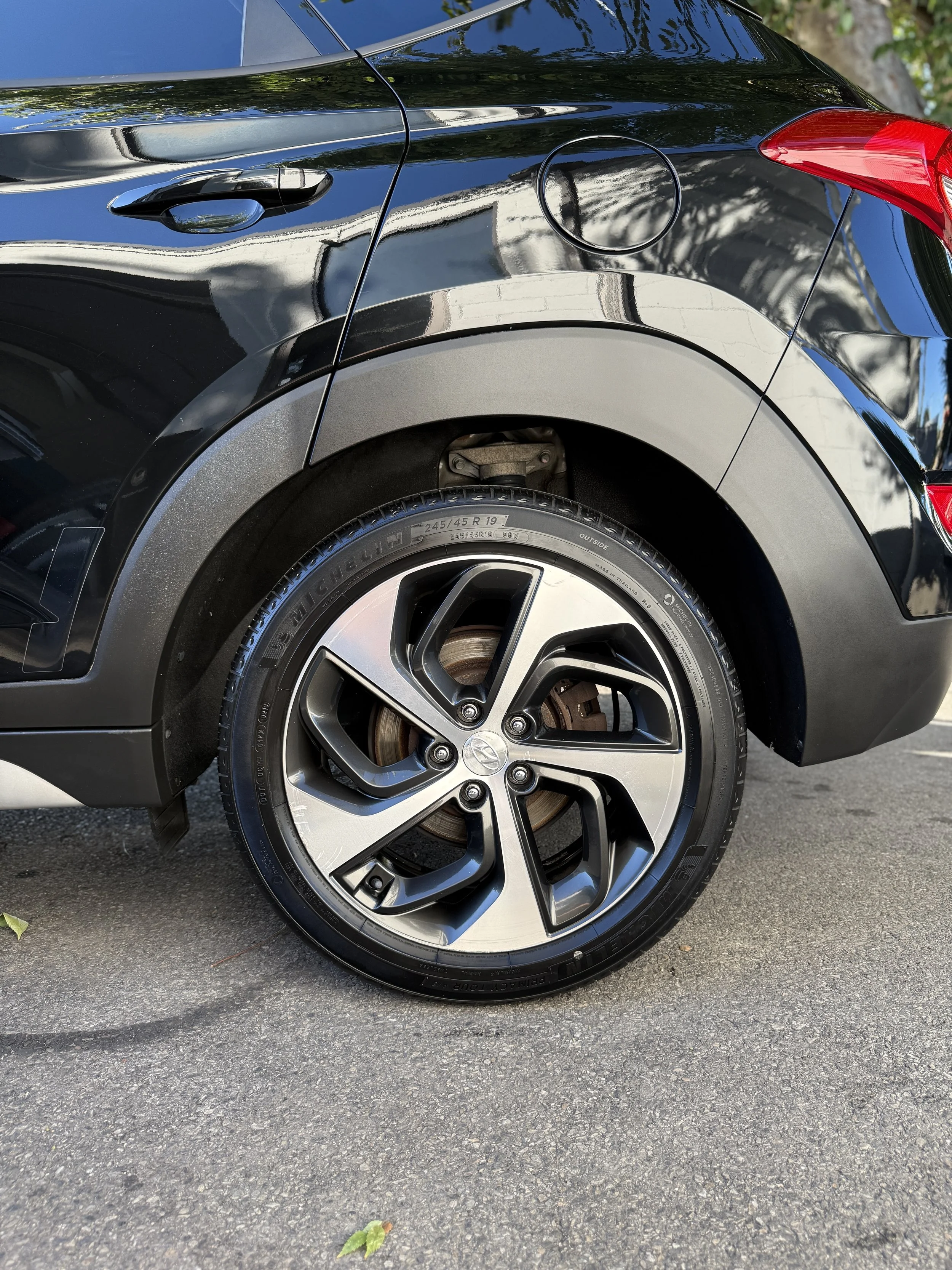Close-up of the rear right side of a black car, showing the wheel, tire, and part of the vehicle's body, with a glimpse of the rear brake and fender.