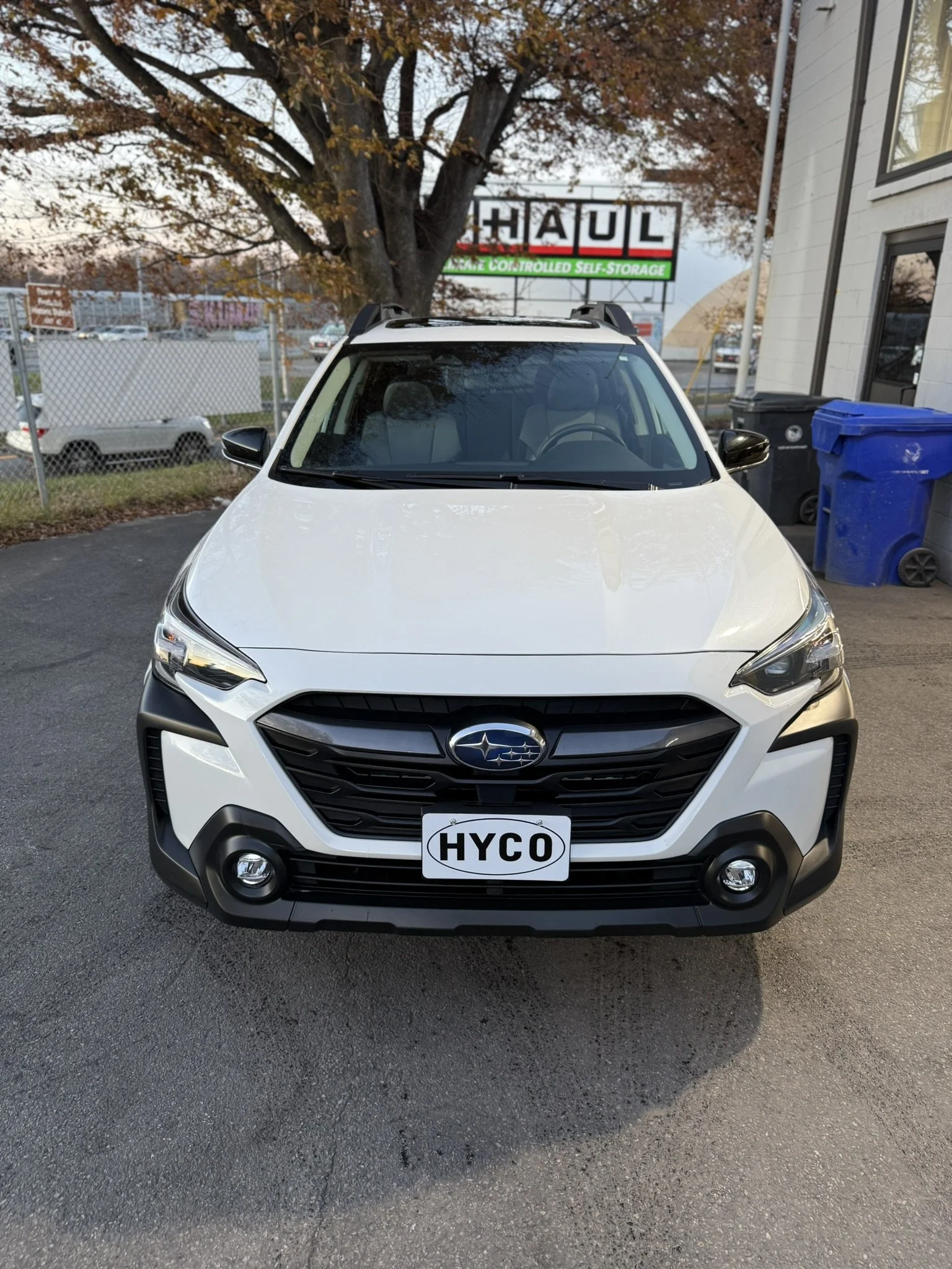White Subaru SUV with custom license plate that says 'HYCO' parked in a lot near a tree and a building, with a U-Haul sign and a fence in the background