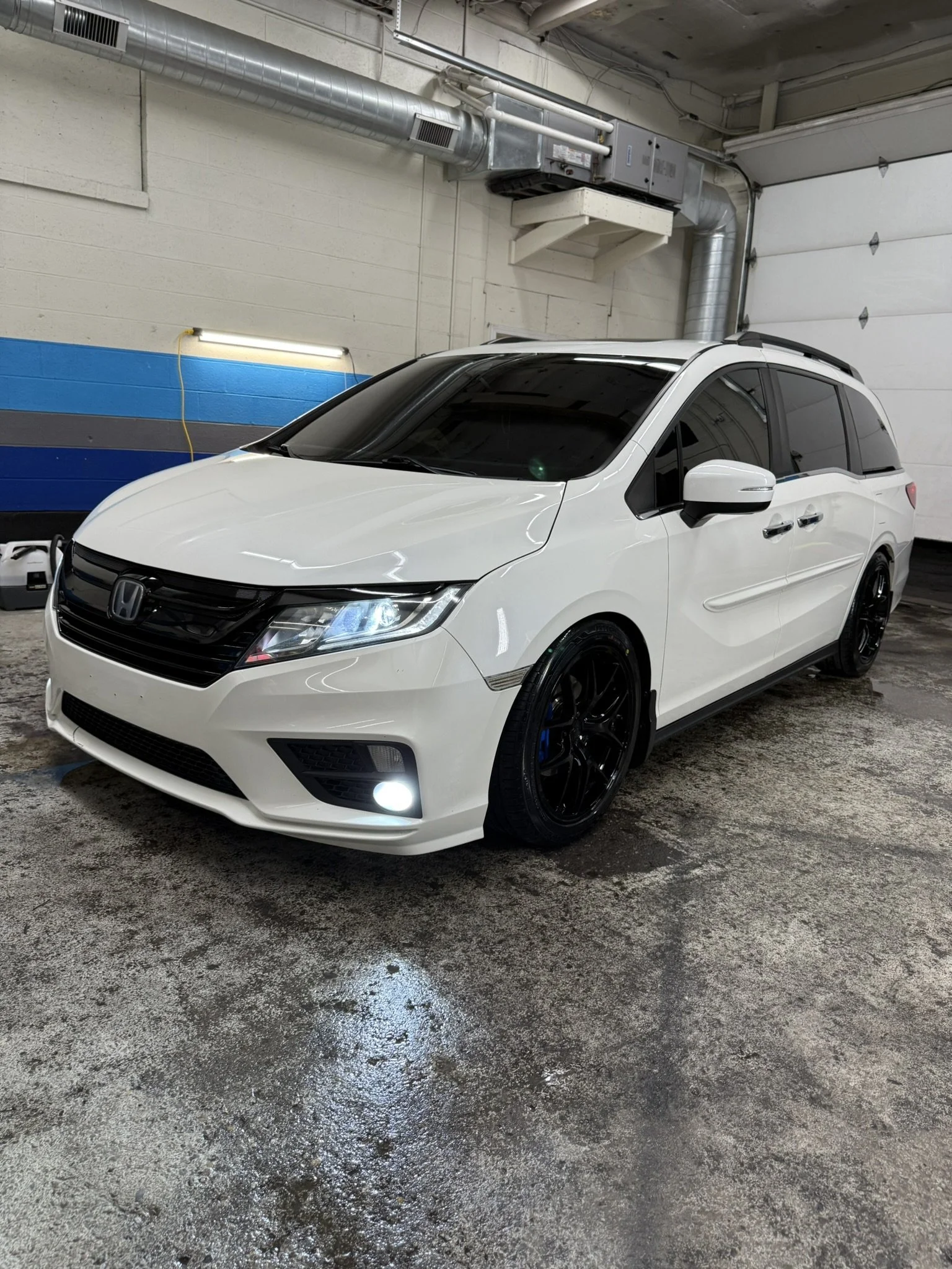 White Honda Odyssey minivan parked inside a garage with wet concrete floor, black wheels, tinted windows, and roof racks.