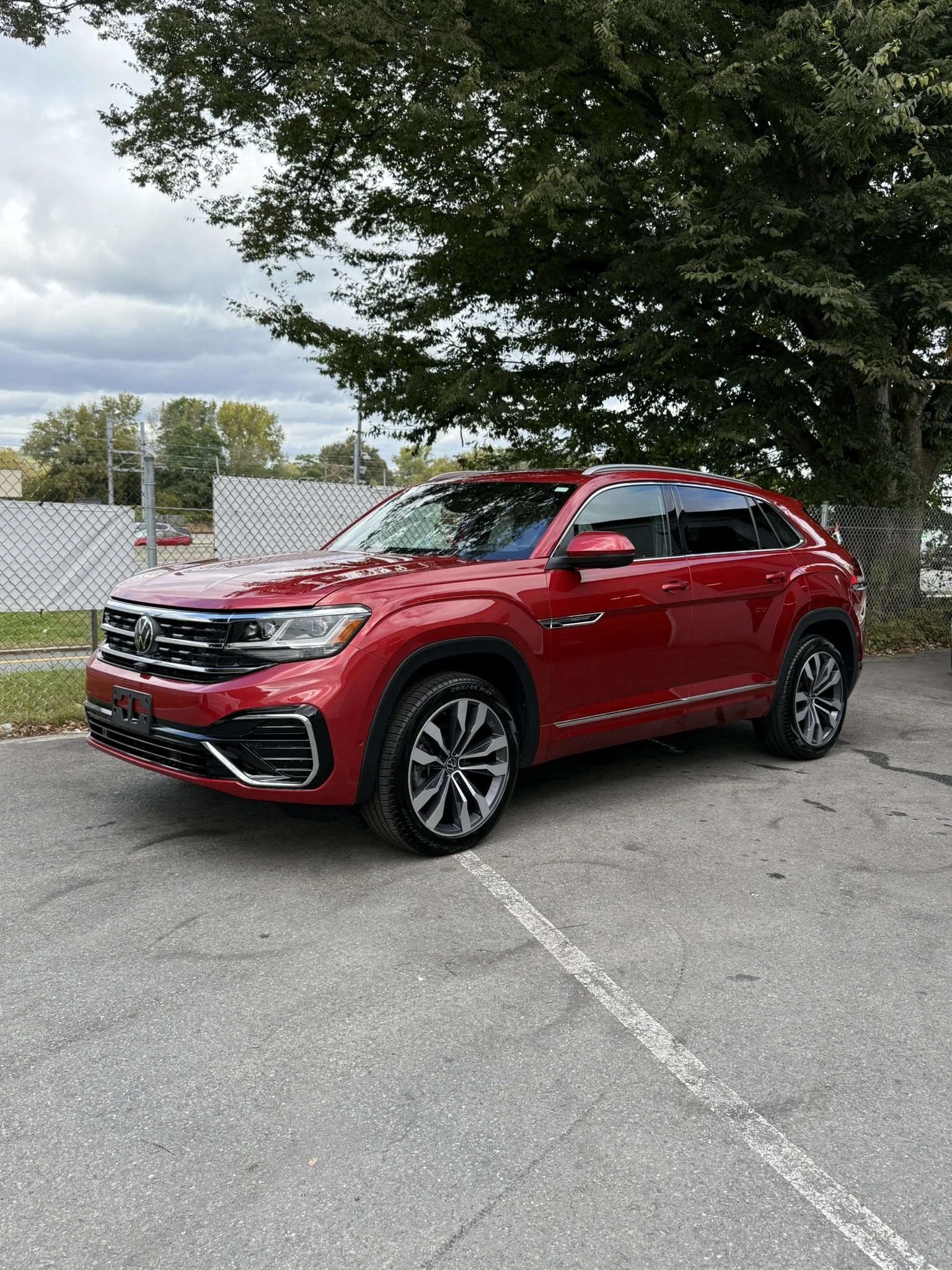 Red Volkswagen SUV parked in a parking lot with a large tree and a chain-link fence in the background under a cloudy sky.