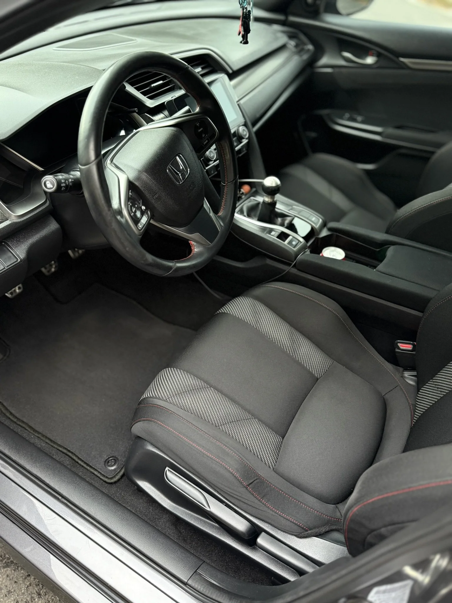 Interior of a Honda car, showing the driver's seat, steering wheel, gear shift, and dashboard with some accessories hanging from the rearview mirror.